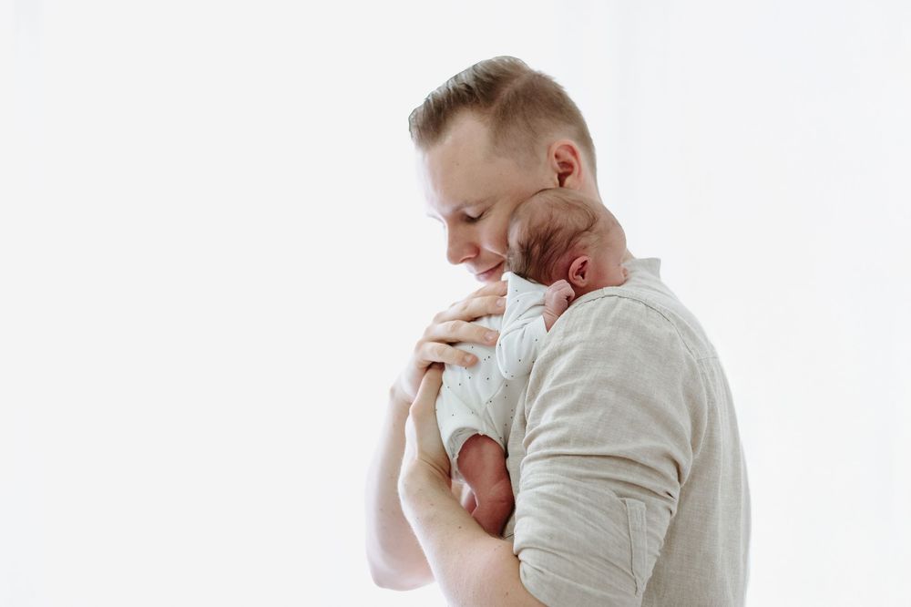 Father tenderly kisses newborn baby on head while cradling infant against chest on white background.