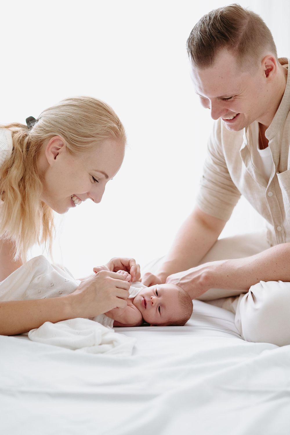 Happy young parents smiling and bonding with their newborn baby lying on a white bed at home.