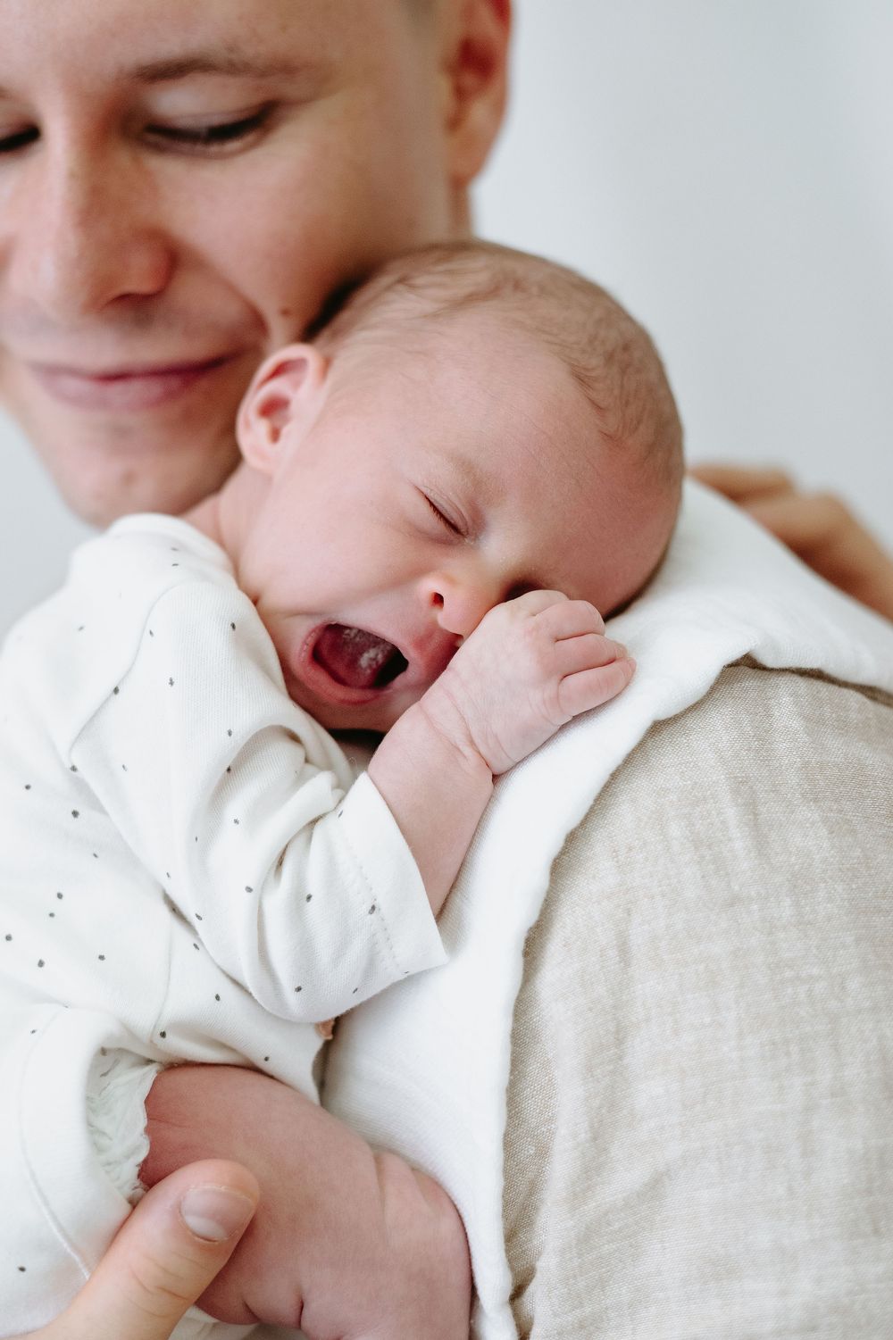 A smiling parent tenderly holds a yawning newborn baby dressed in a white dotted onesie against their chest.