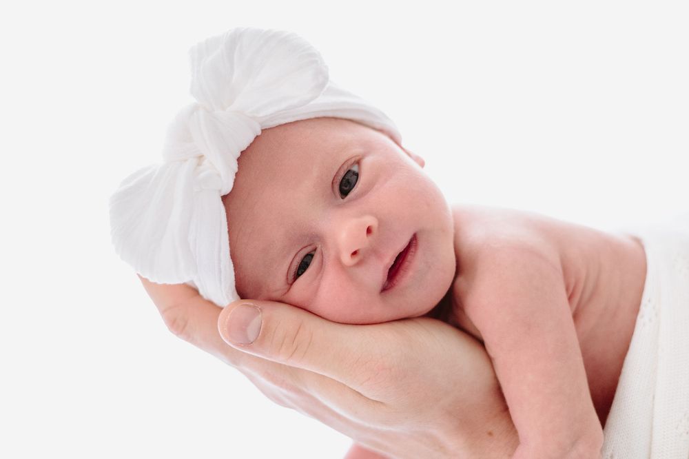 Newborn baby girl wearing a white bow headband, cradled gently in adult hands against a white background.