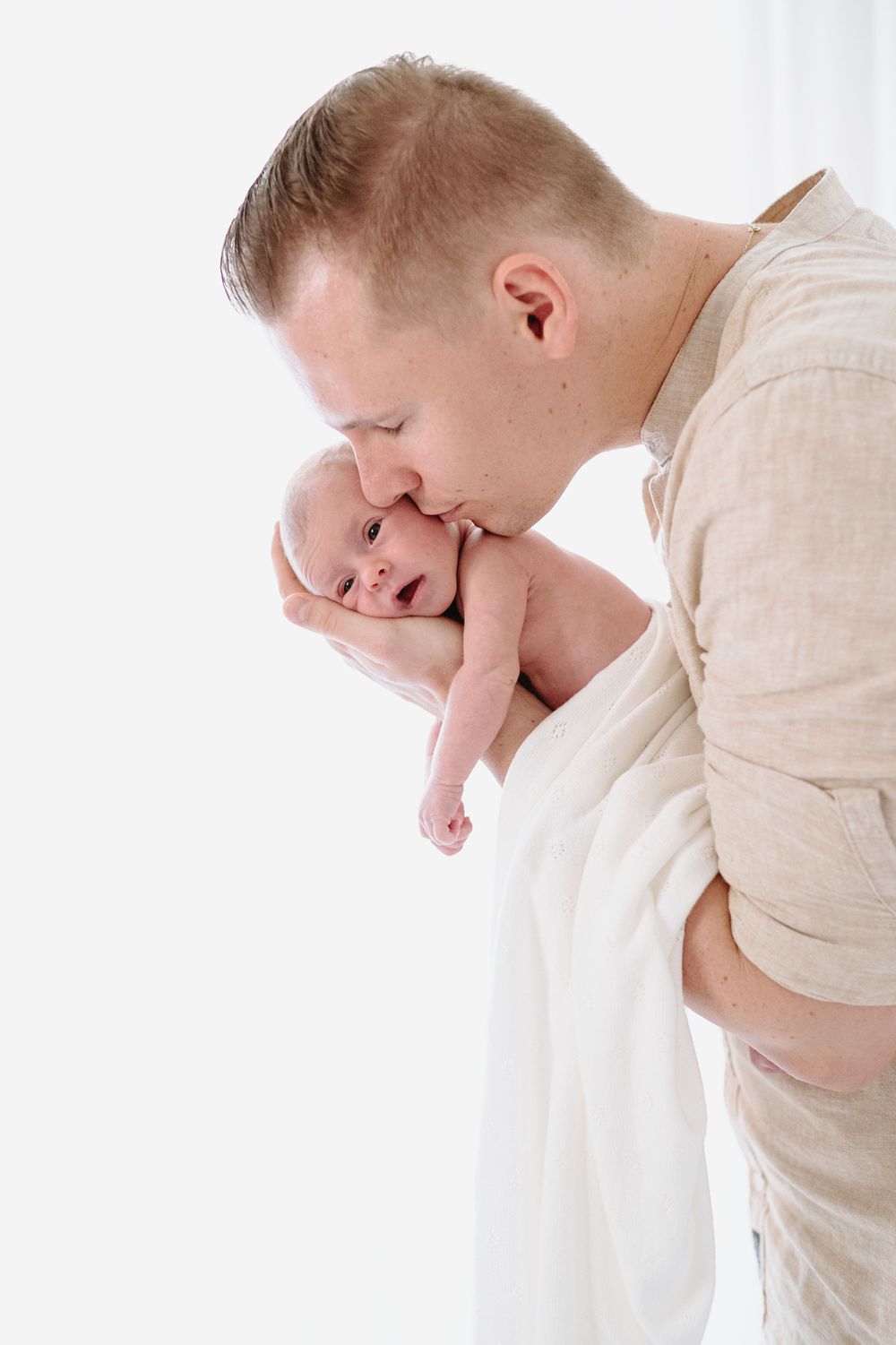 A loving father gently kisses his newborn baby wrapped in a white blanket against a bright white background.