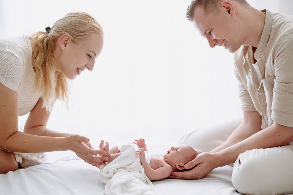 Two loving parents smiling and bonding with their newborn baby lying on a white bed.