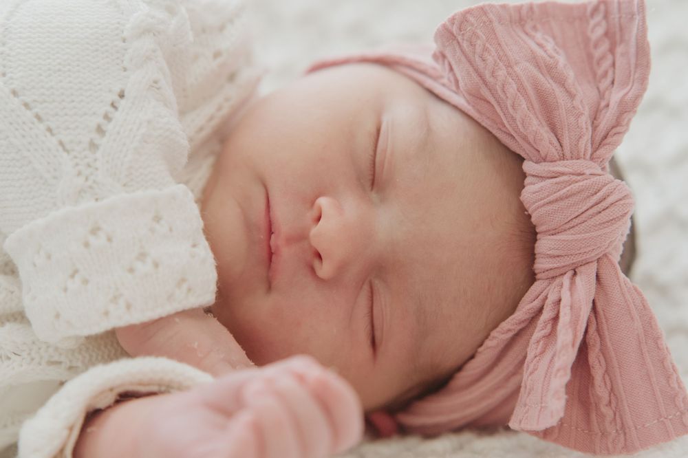 Peaceful newborn baby girl sleeping in a pink bow headband and white knit outfit, soft natural light photography.