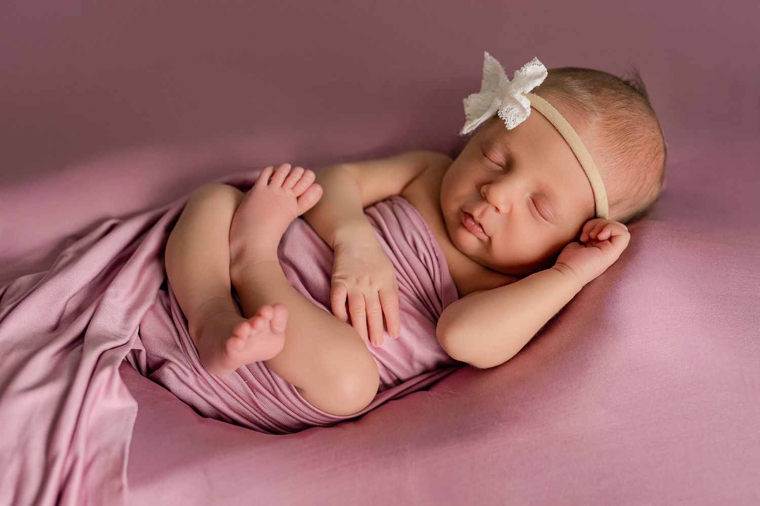 Sleeping newborn in red wrap wearing floral bow headband on white fur backdrop.