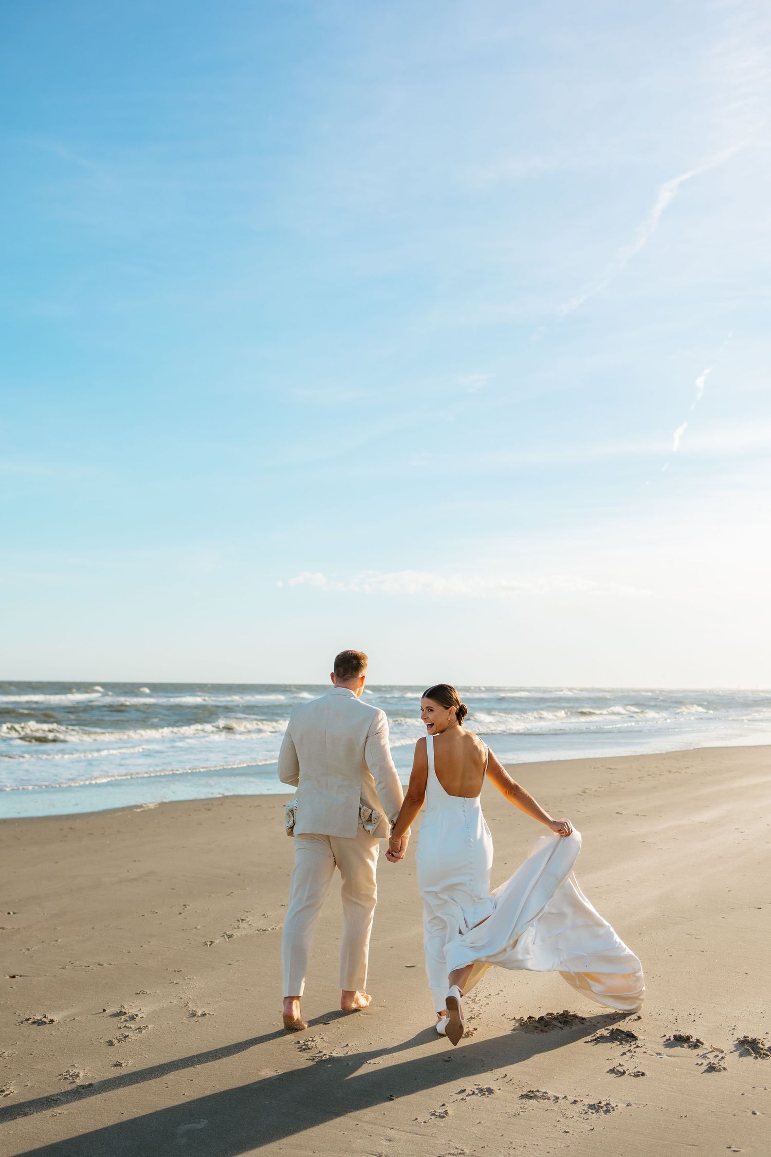 A couple in wedding attire walks along a sunny beach shoreline with waves crashing in the background.