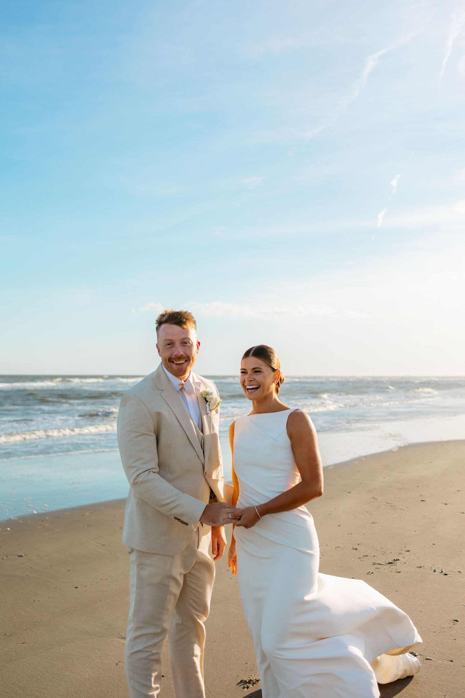 A happy couple shares a joyful moment on a sunny beach during their wedding photoshoot at sunset.