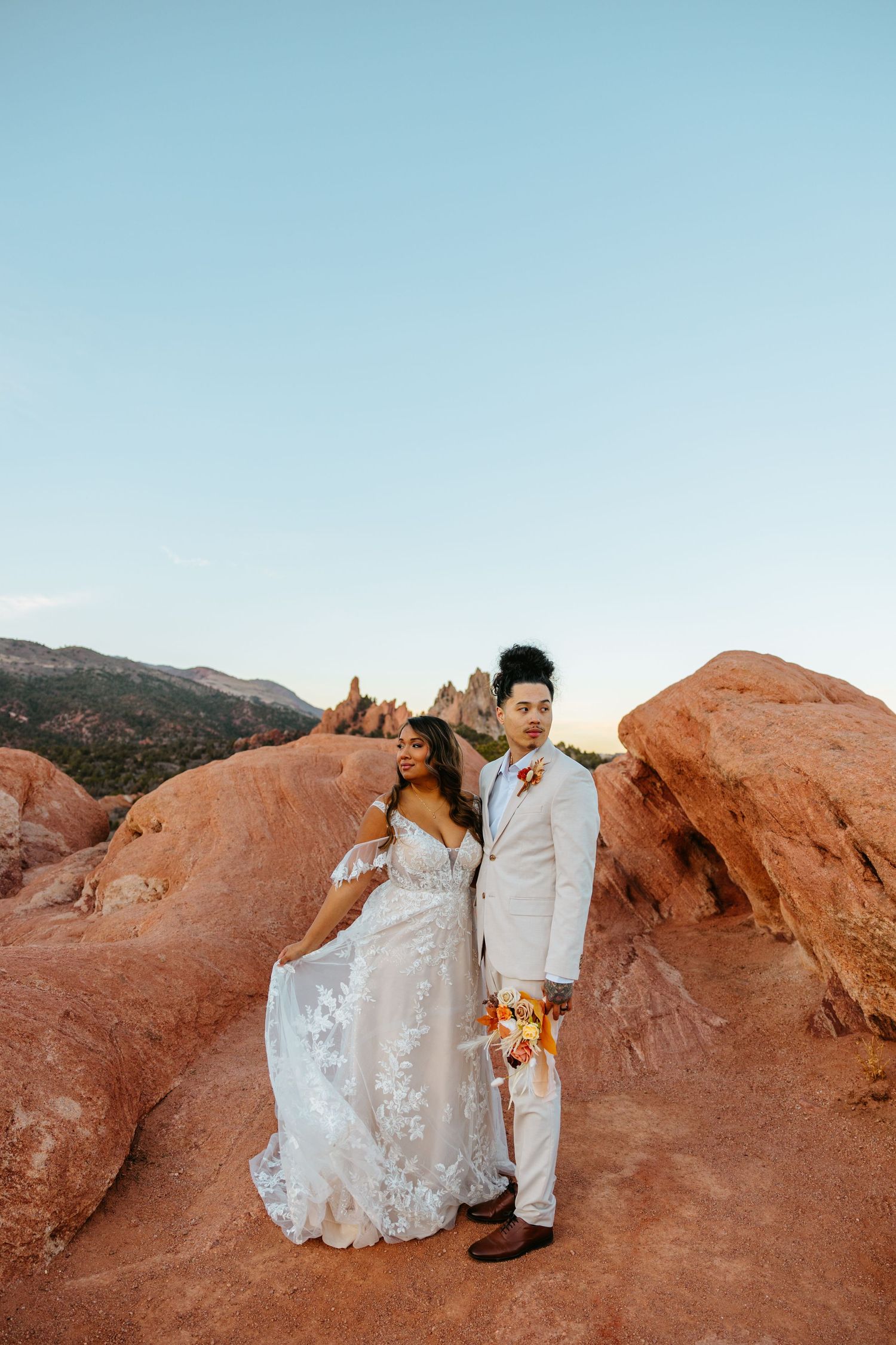 Wedding couple poses with their dog against stunning red rock formations in desert landscape.