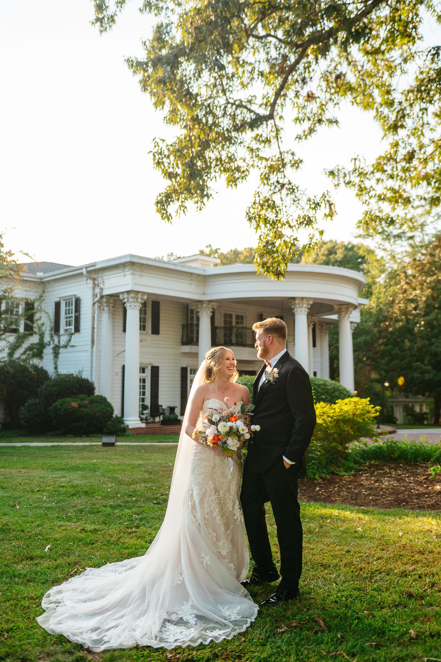 Couple in wedding attire stands on a sunny lawn outside a stately white mansion with columns.
