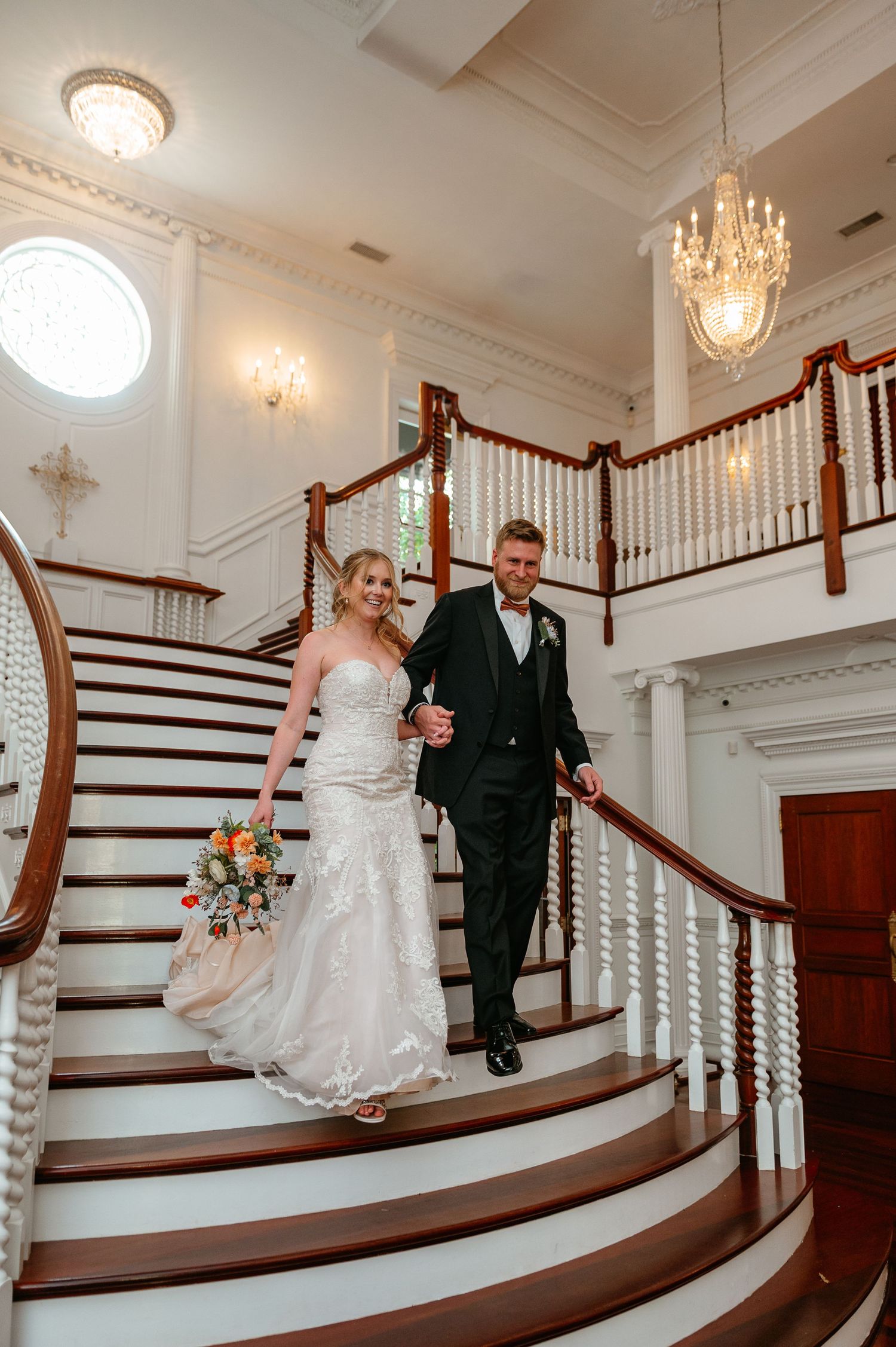 A couple descends an elegant white staircase with chandelier during a wedding at a historic venue.