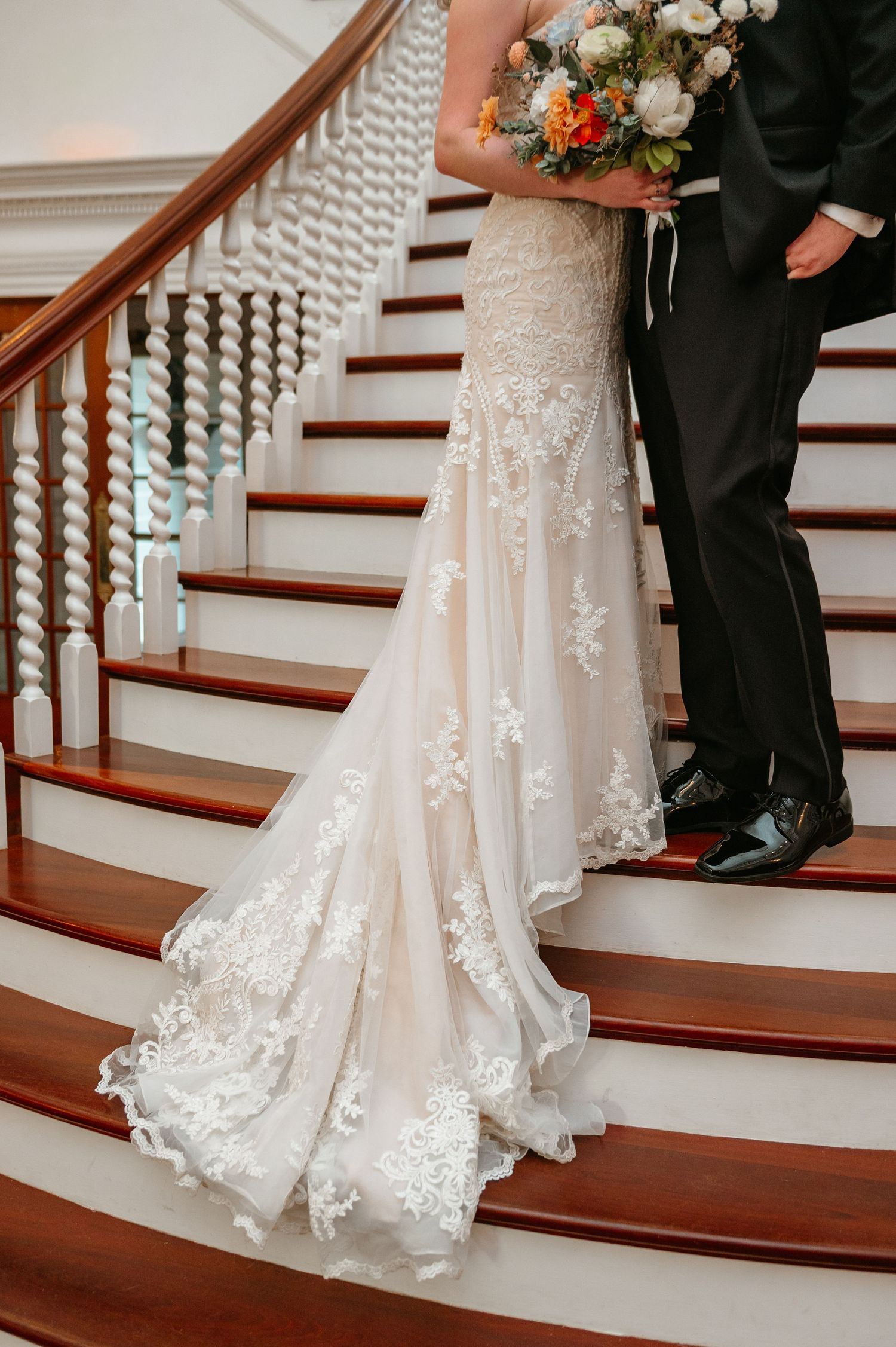 Close-up of intricate lace wedding dress train cascading down wooden stairs with floral bouquet details.
