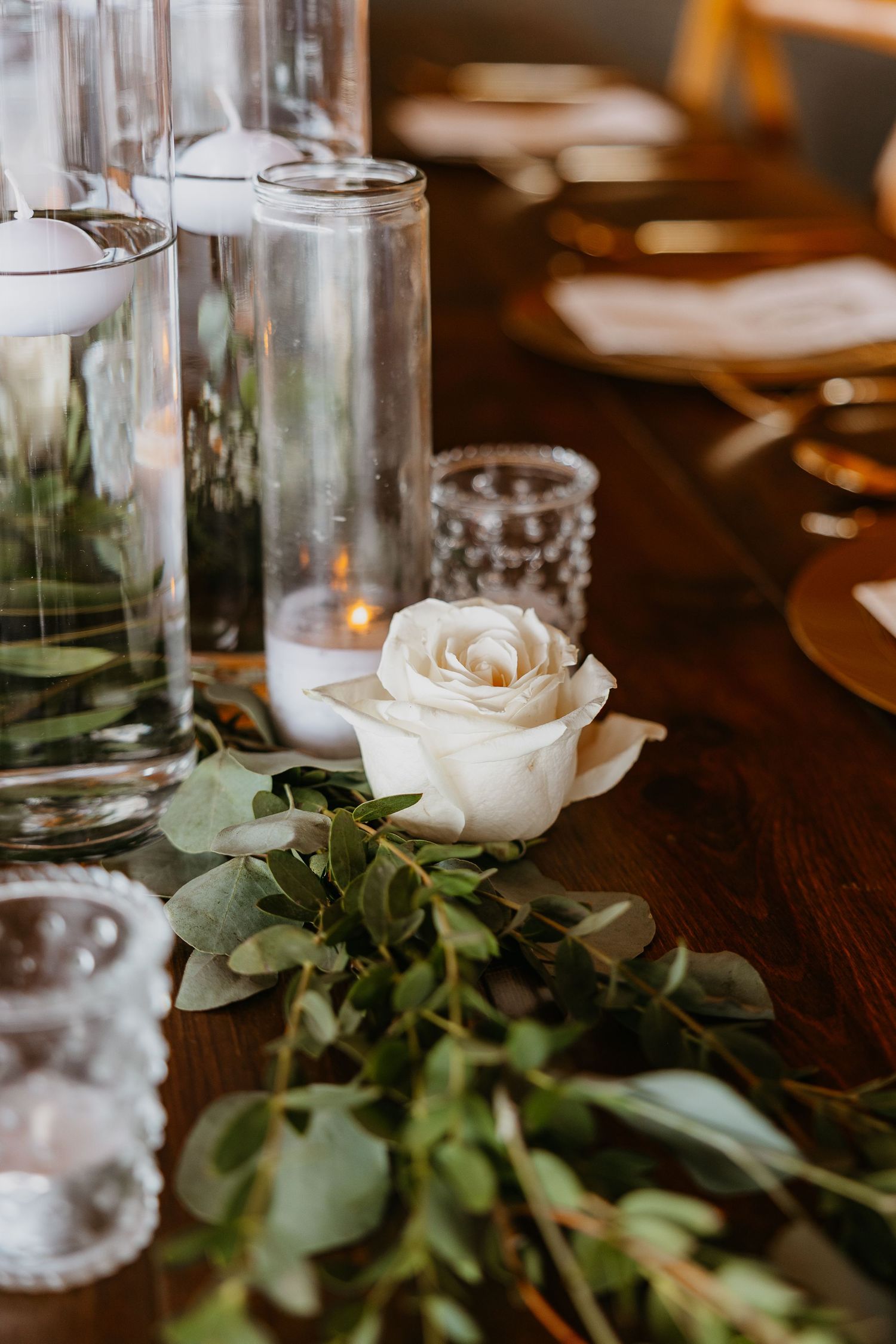 Fresh white roses and greenery arranged on a dark wooden table with glass vases creating an elegant table setting.