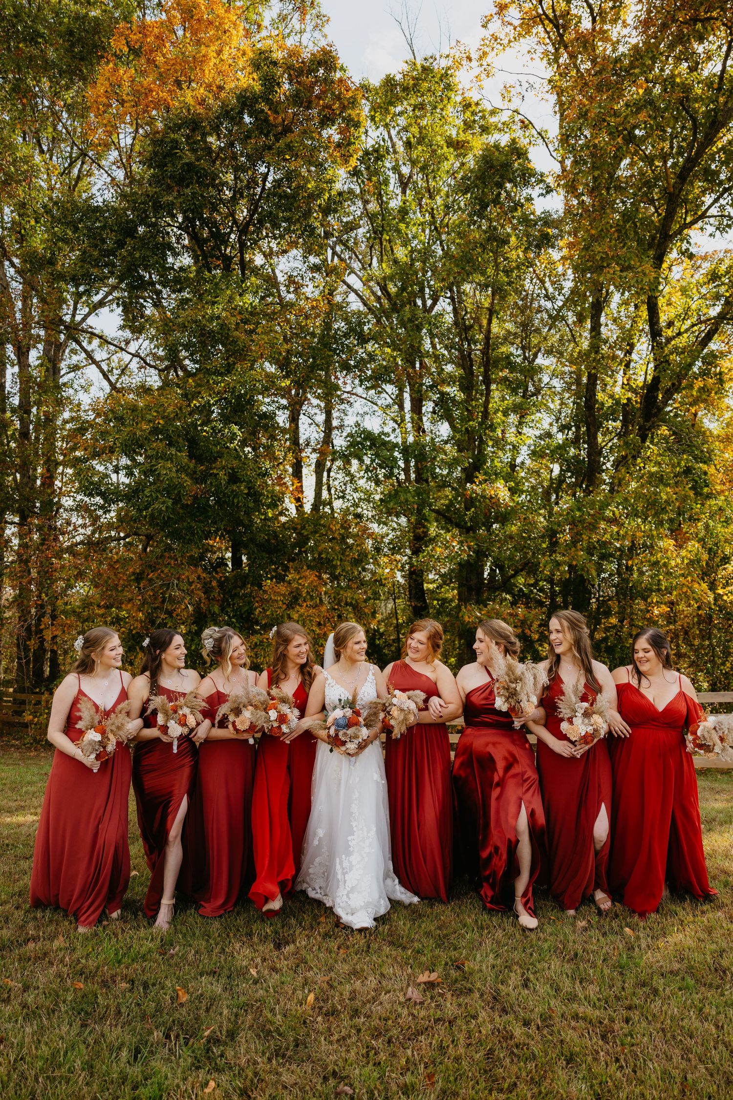Bridal party in flowing red dresses poses together on autumn grass during sunset.