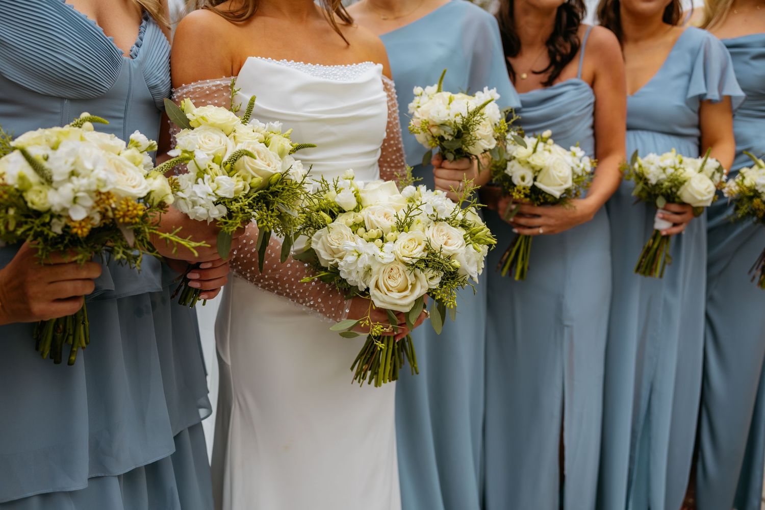 Close-up of wedding bouquets featuring white roses held by bridesmaids in dusty blue dresses.