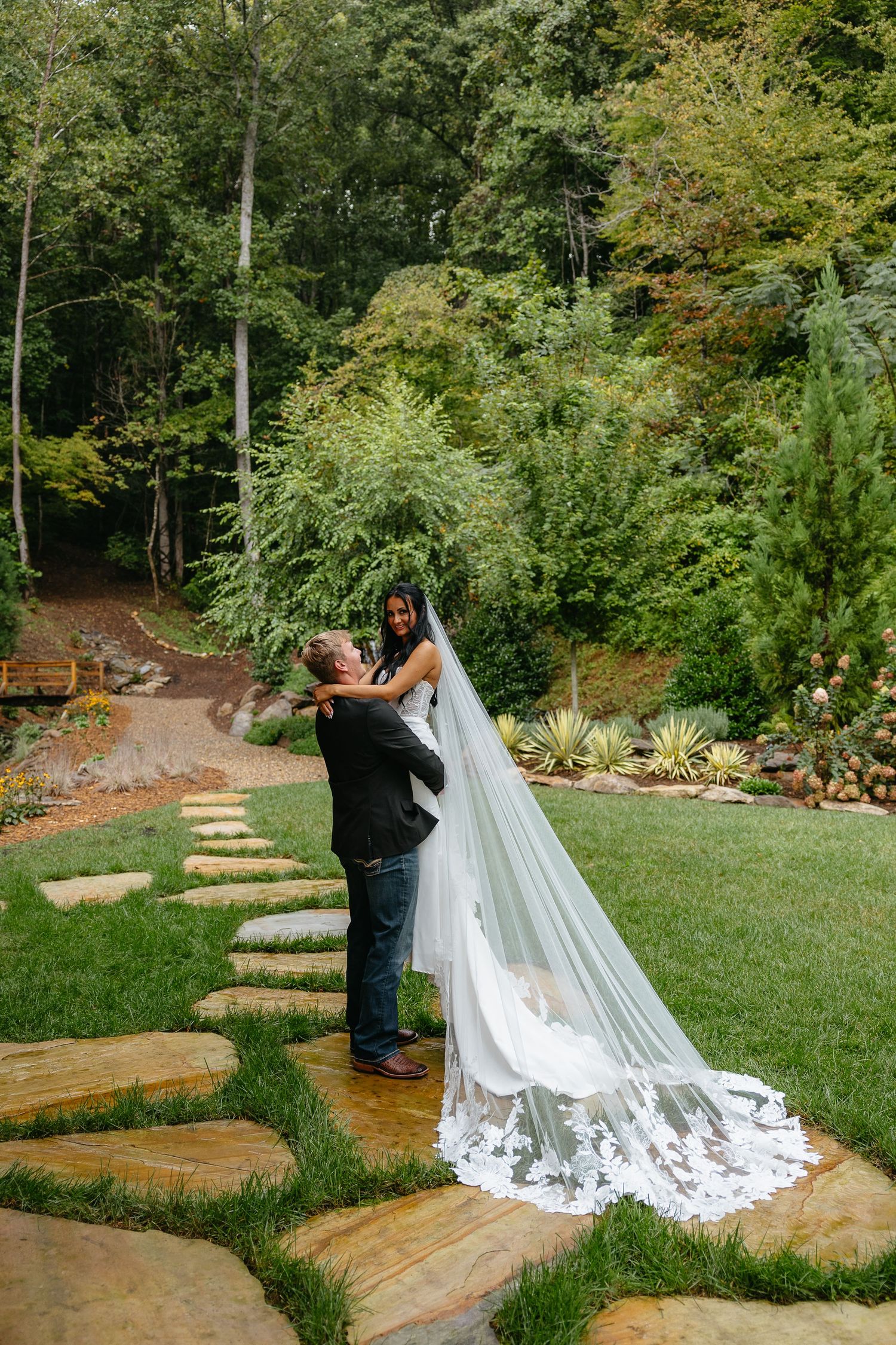 A scenic outdoor wedding photography session showing a long flowing white veil trailing on stone steps in a garden setting.