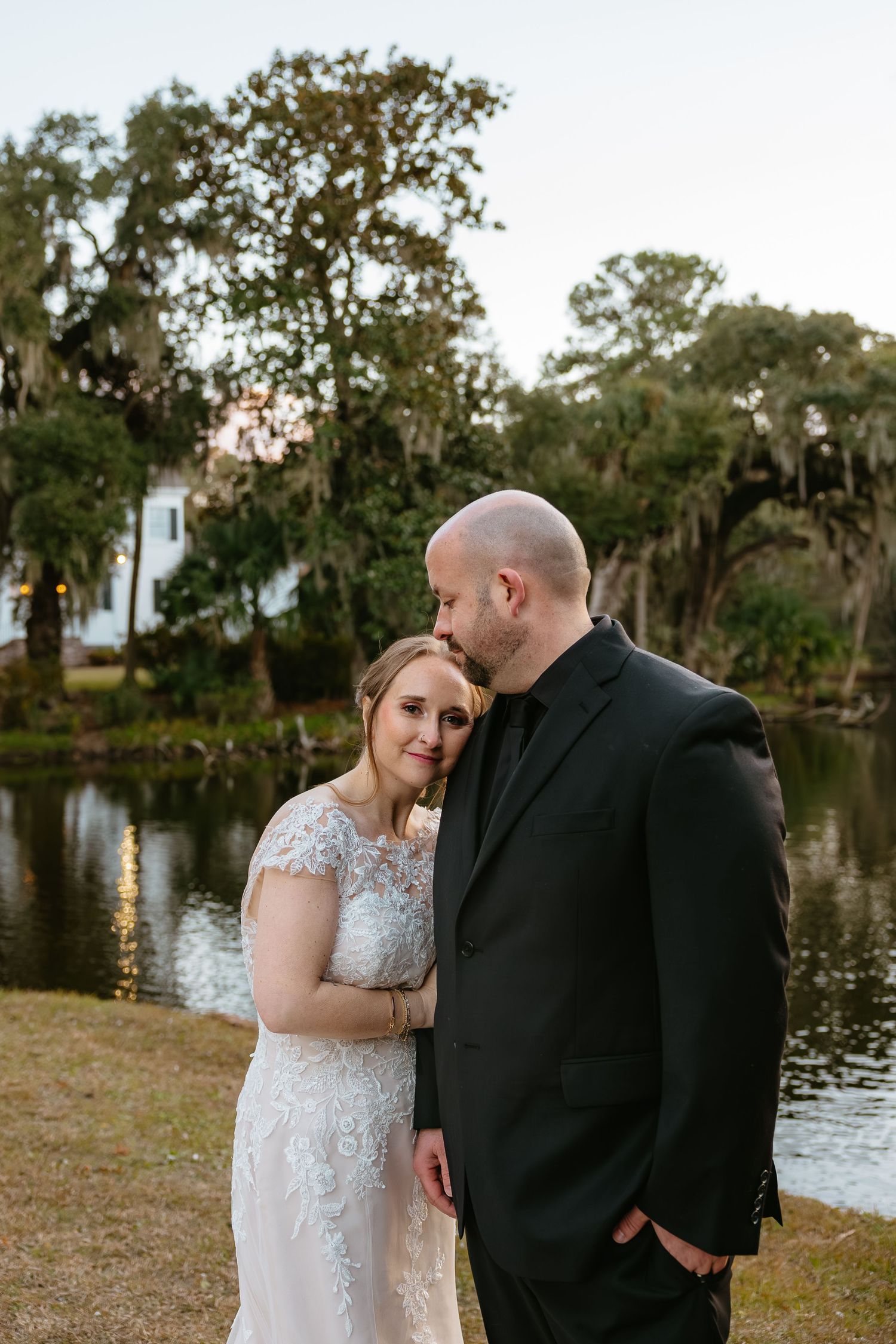 A romantic couple in formal attire shares intimate moments by a serene lake with Spanish moss-draped trees in the background.