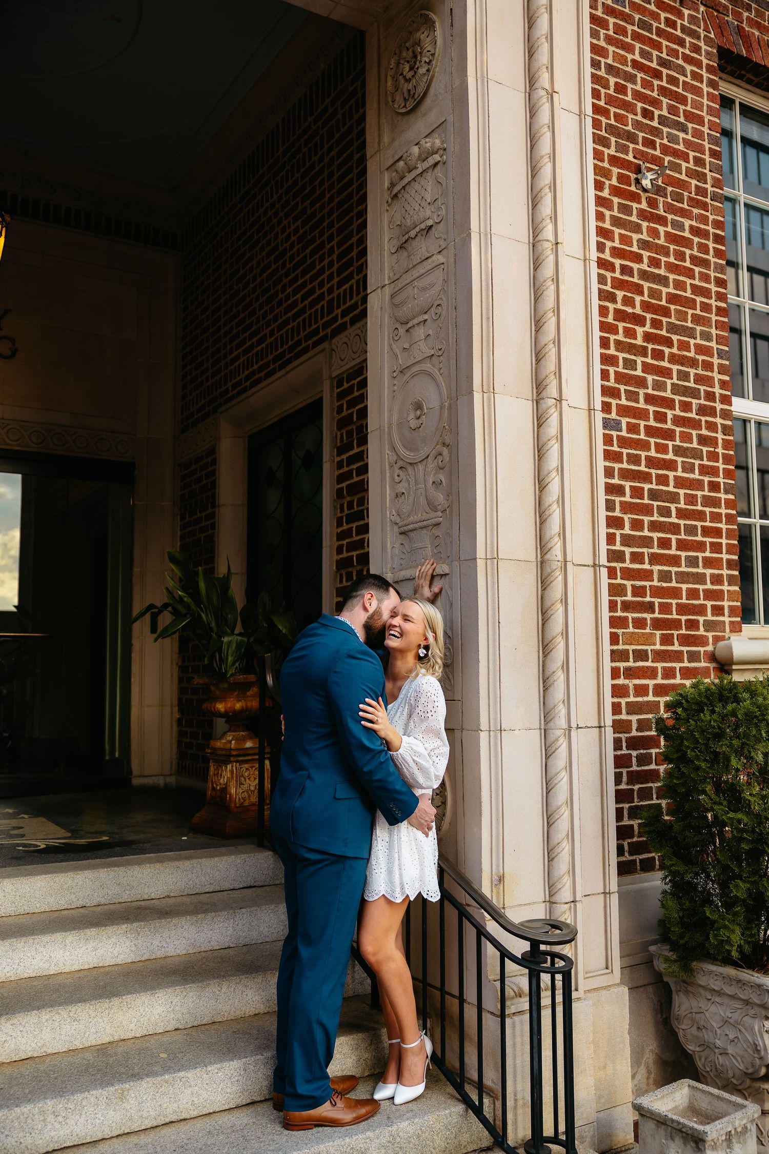 A couple shares an intimate moment embracing on a brick building's front steps in the afternoon light.