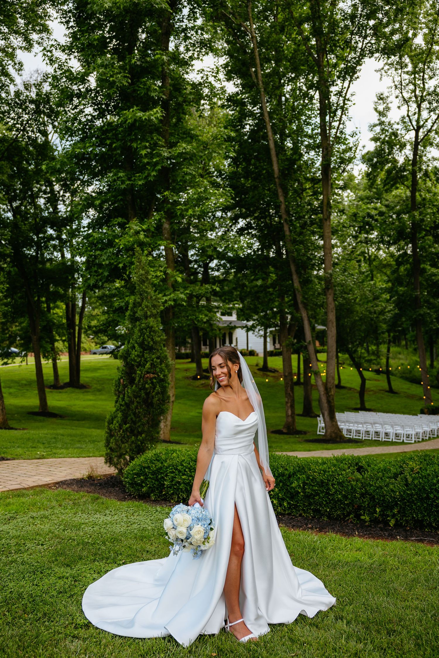 A bride in an elegant white wedding dress poses outdoors in a lush garden setting with trees and white chairs arranged.