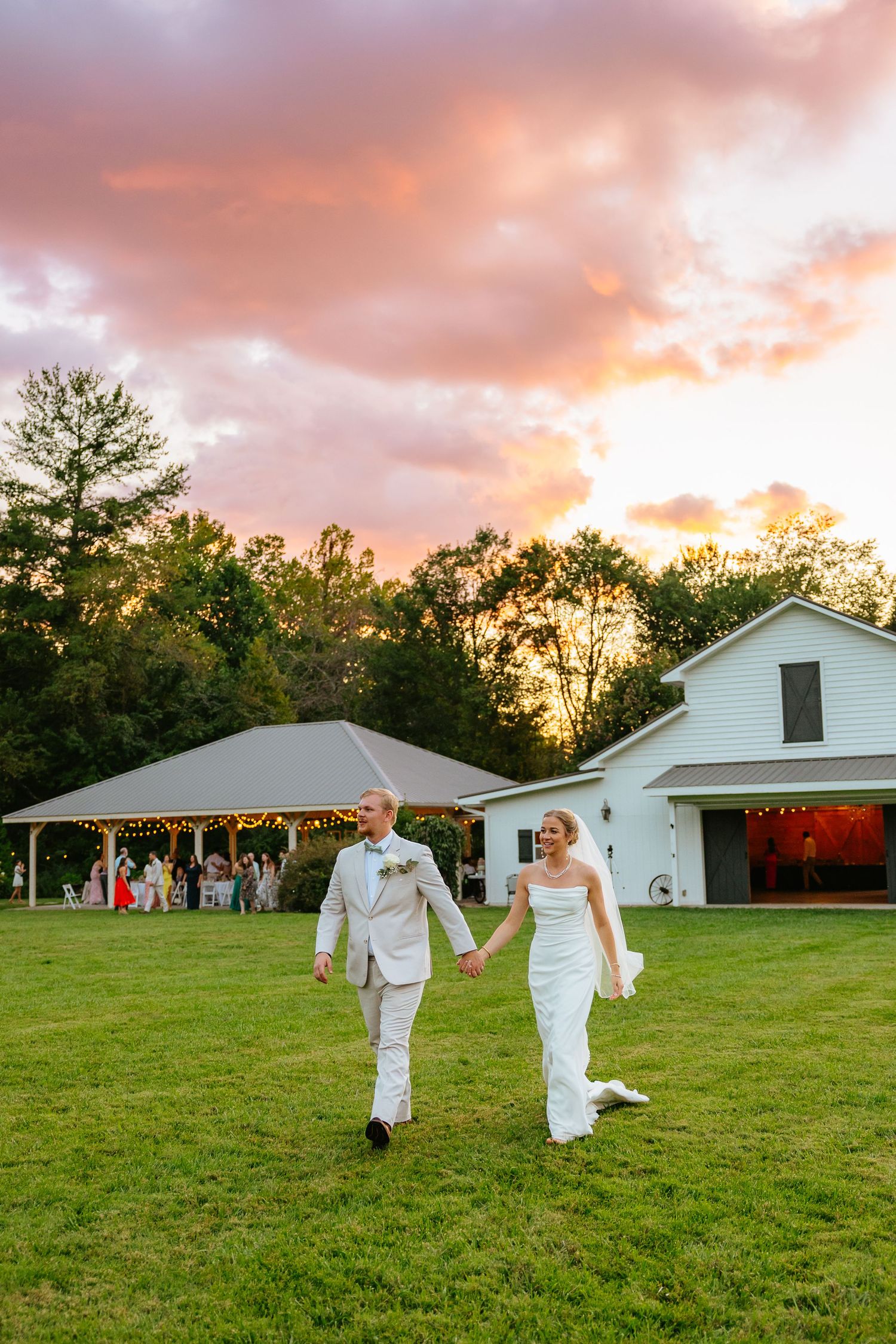 A couple in white attire walks across a grassy field at sunset with a rustic white building in the background.