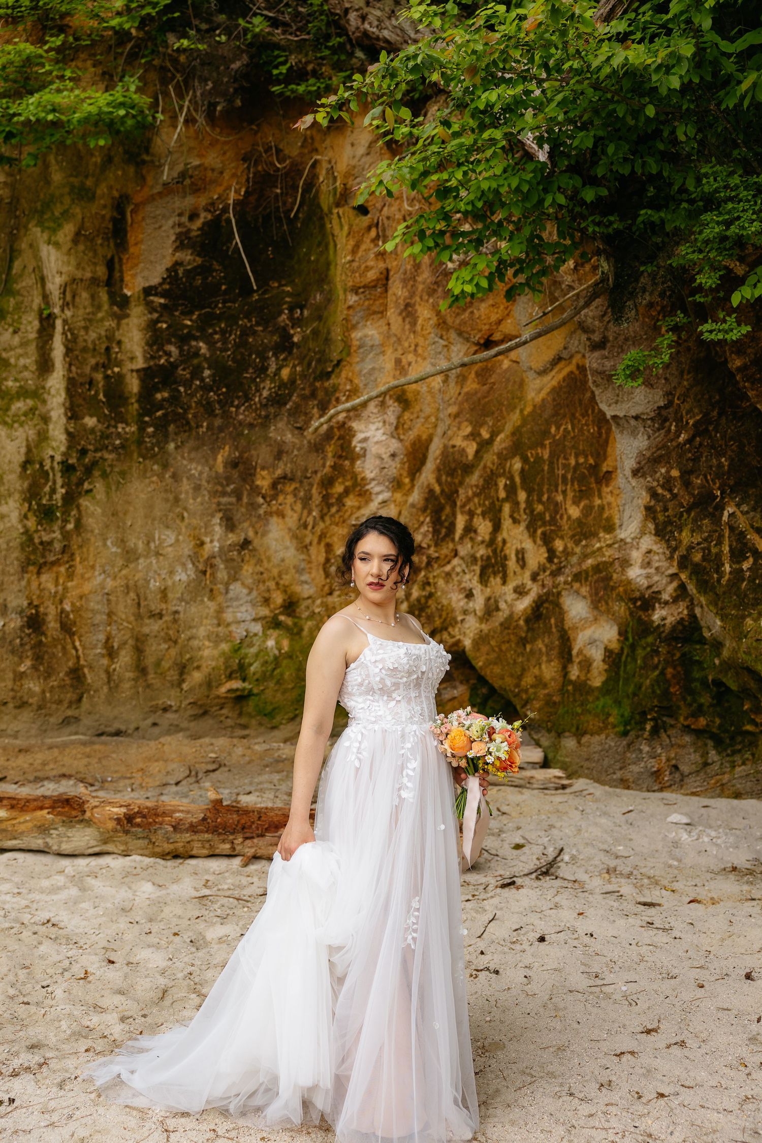 Elegant bride in flowing white dress poses against rocky cliff wall in natural outdoor setting.