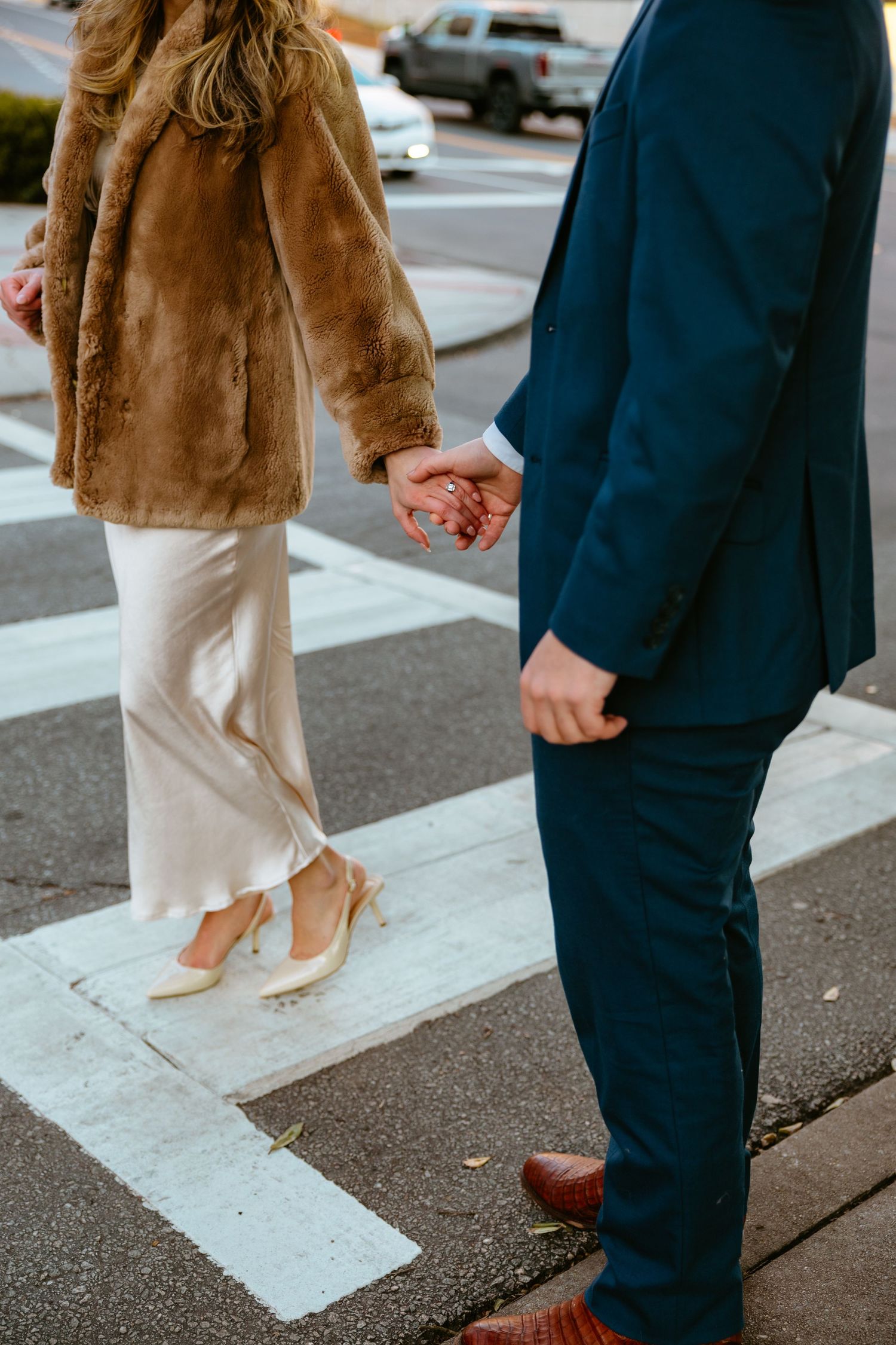 A couple holding hands on a crosswalk, wearing a brown fur coat and navy suit with brown leather shoes.