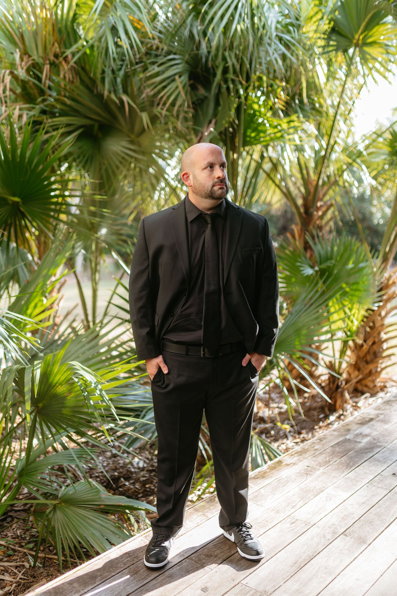 A person in all black attire stands on a wooden deck surrounded by tropical palm trees and lush greenery.