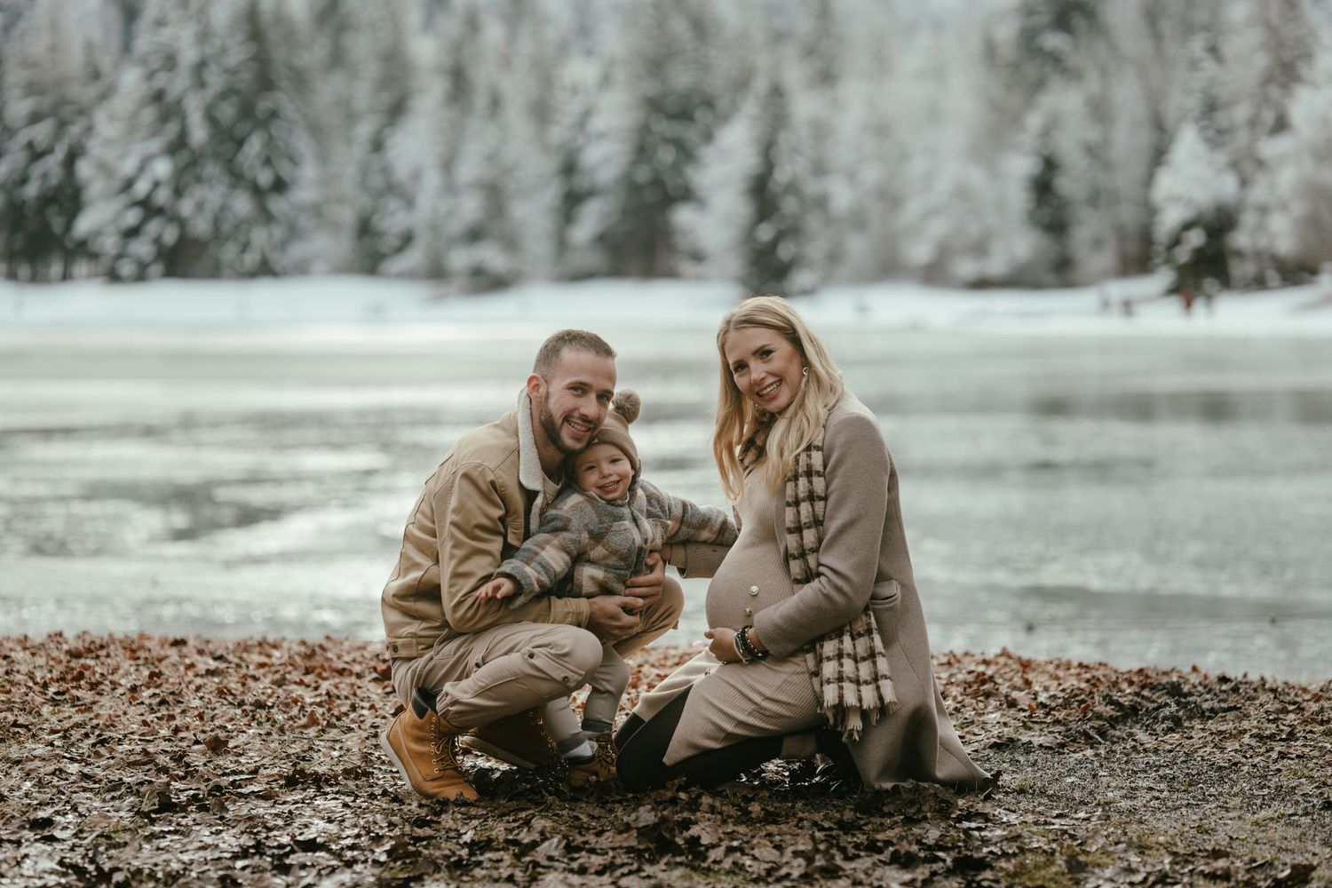 Winter engagement photo of couple with dog by frozen lake surrounded by snow-covered trees.
