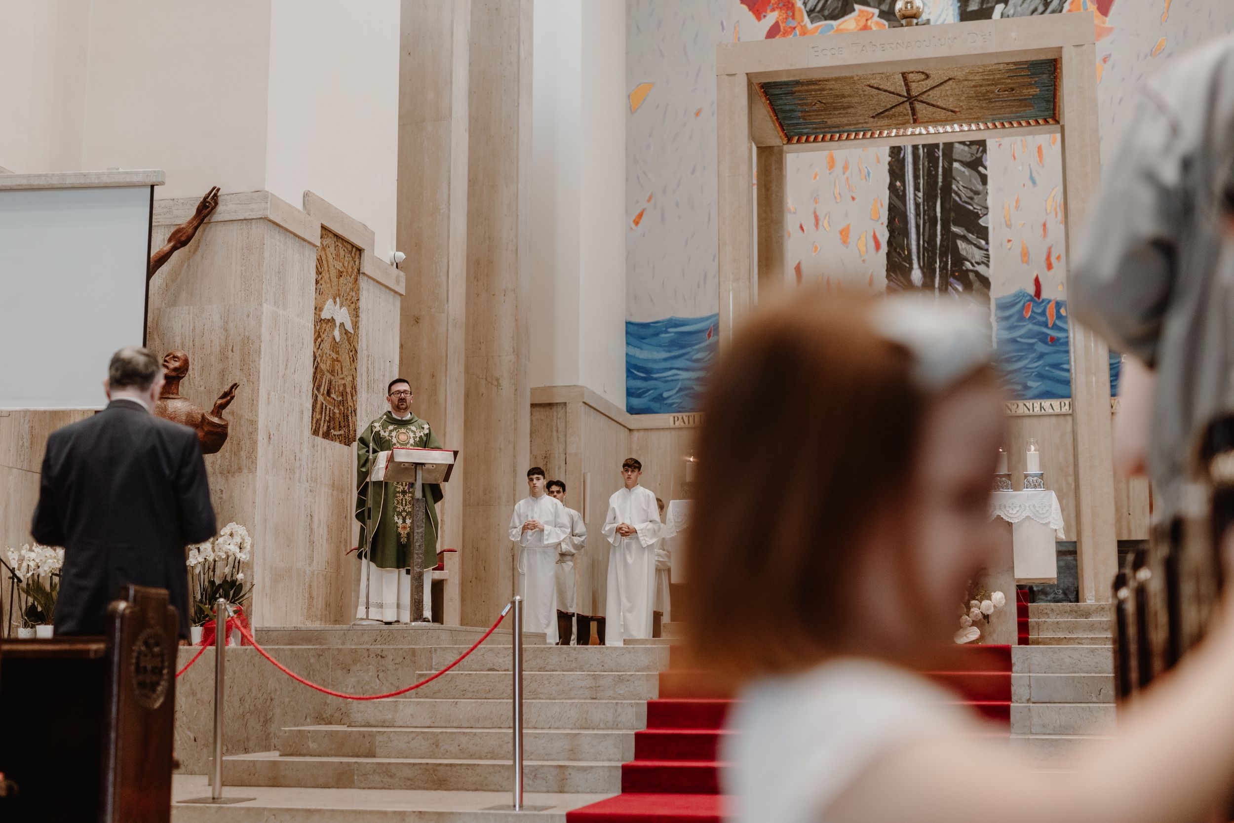 Front view of a child out of focus, with the priest in the background at the altar delivering a sermon.