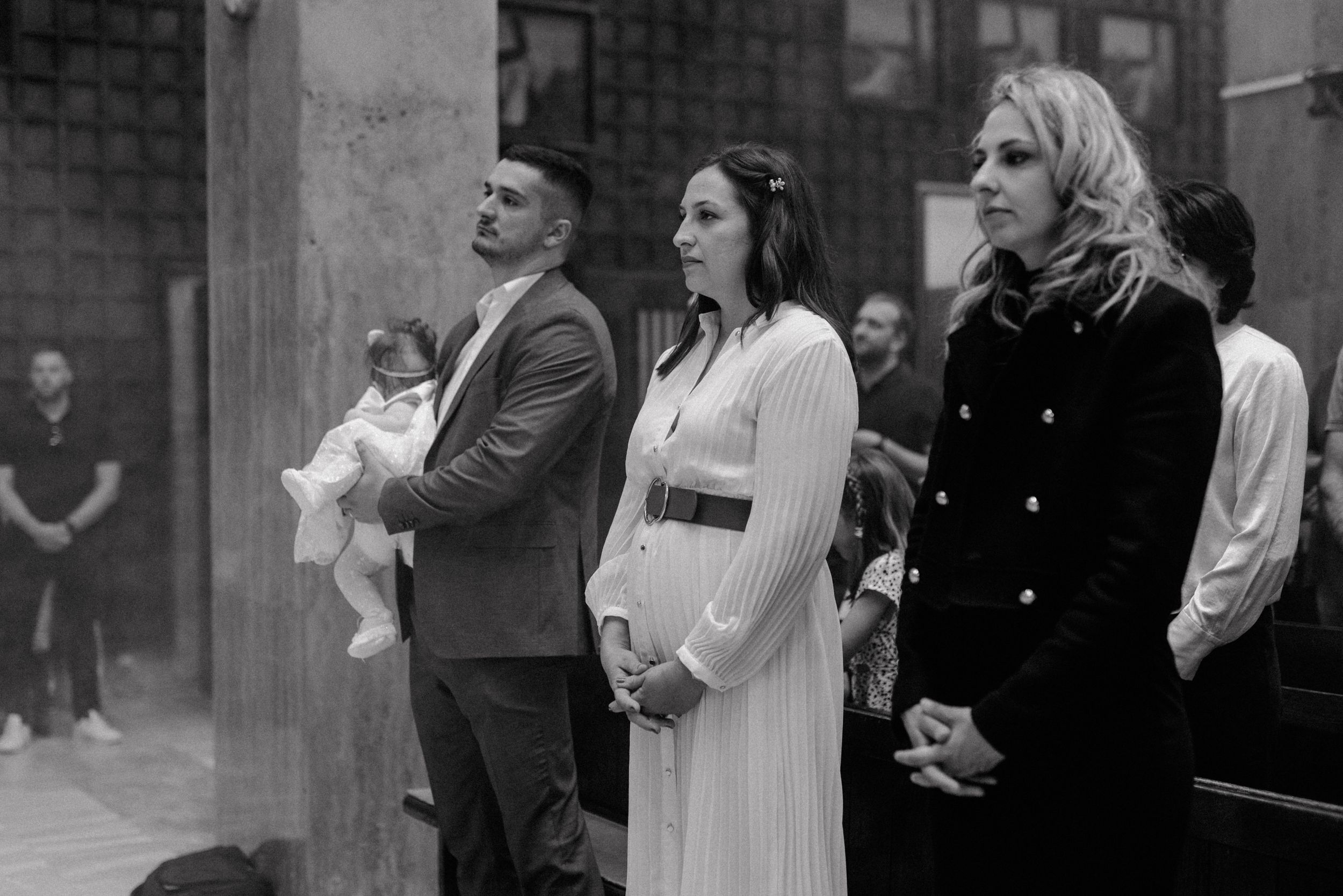 Father holding his daughter during the christening ceremony, with the mother and godmother standing alongside them.