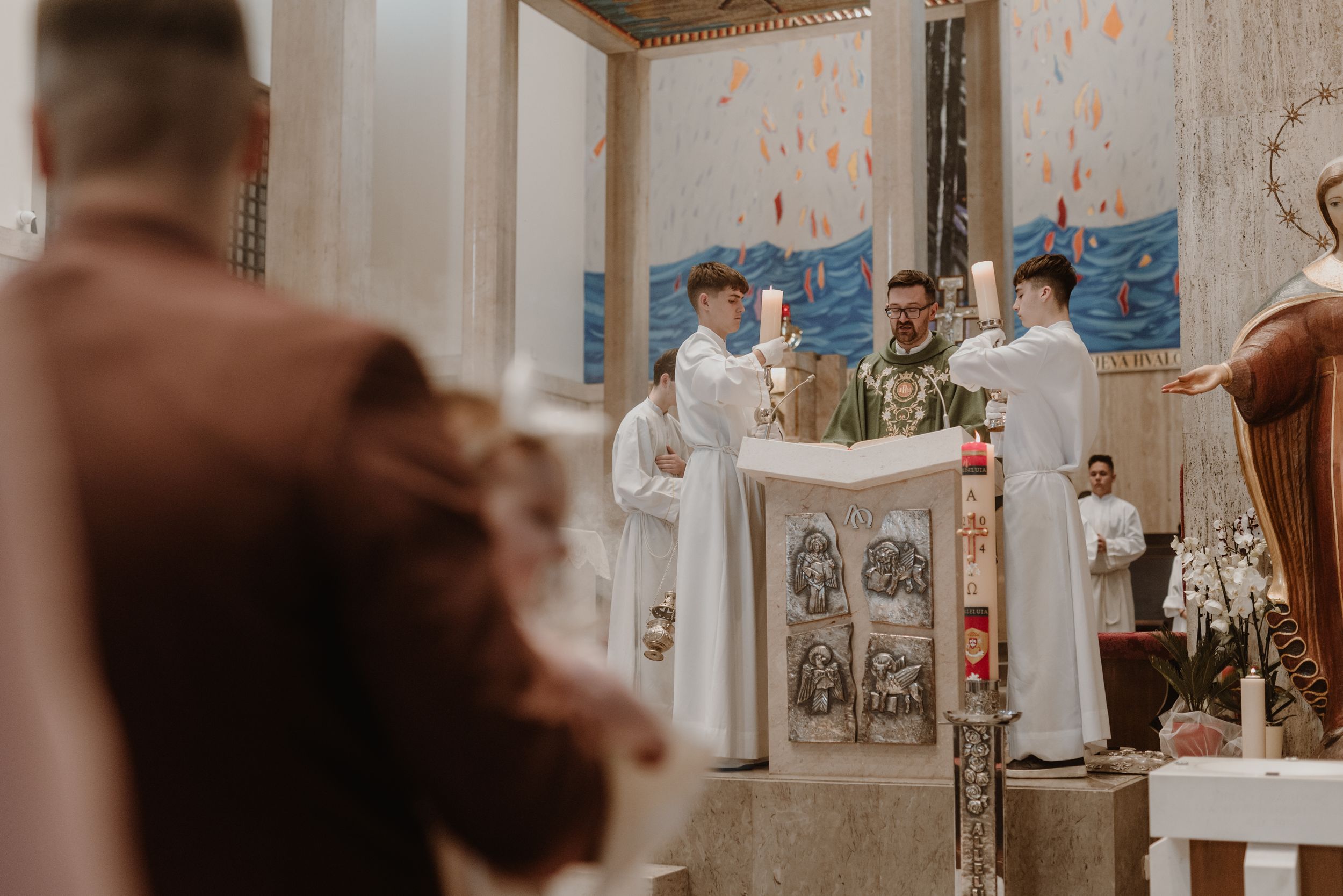 Father and his child out of focus in the foreground, while the priest and altar boys holding candles.