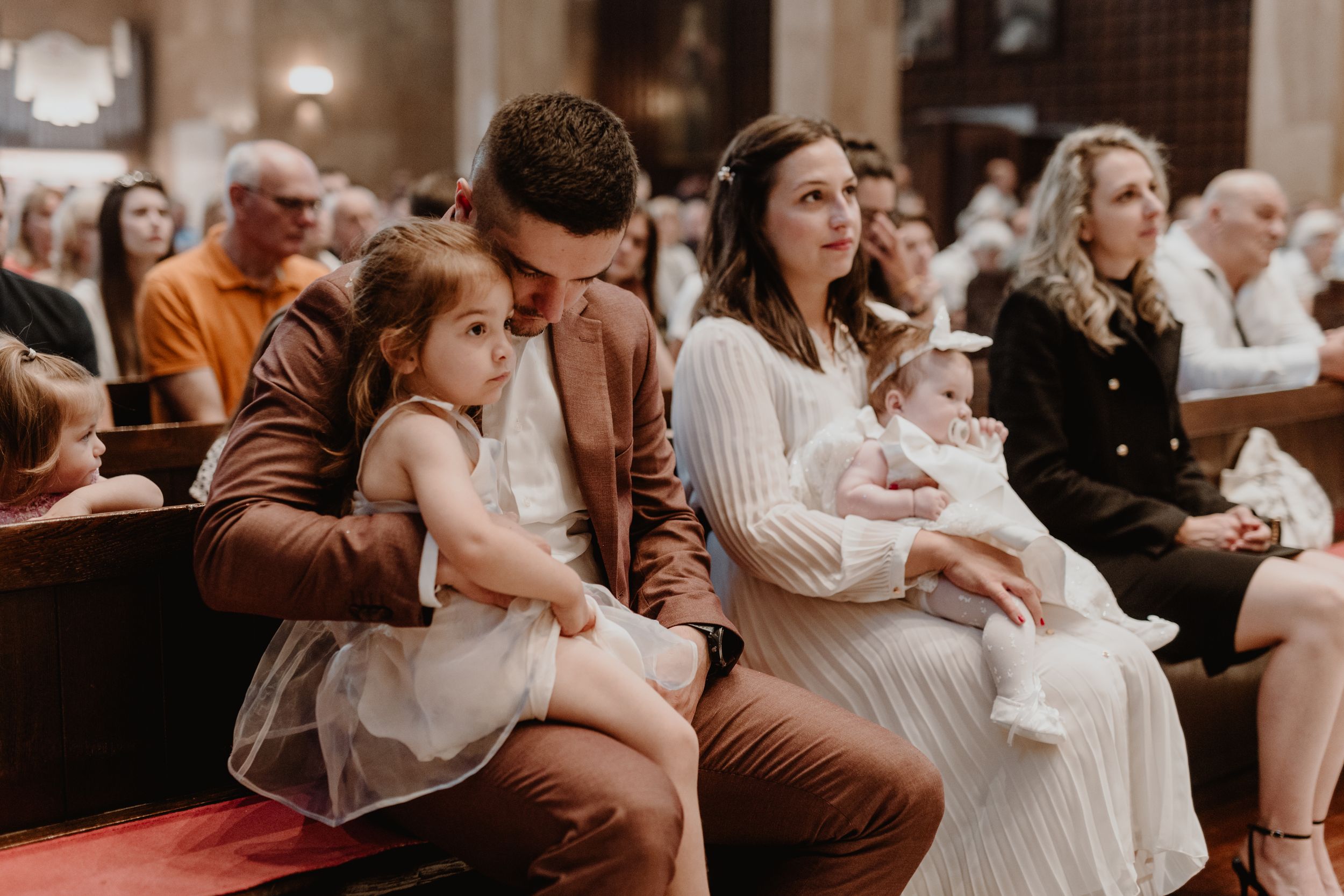 Dad holding his older daughter while mom holds their younger daughter during the christening ceremony.