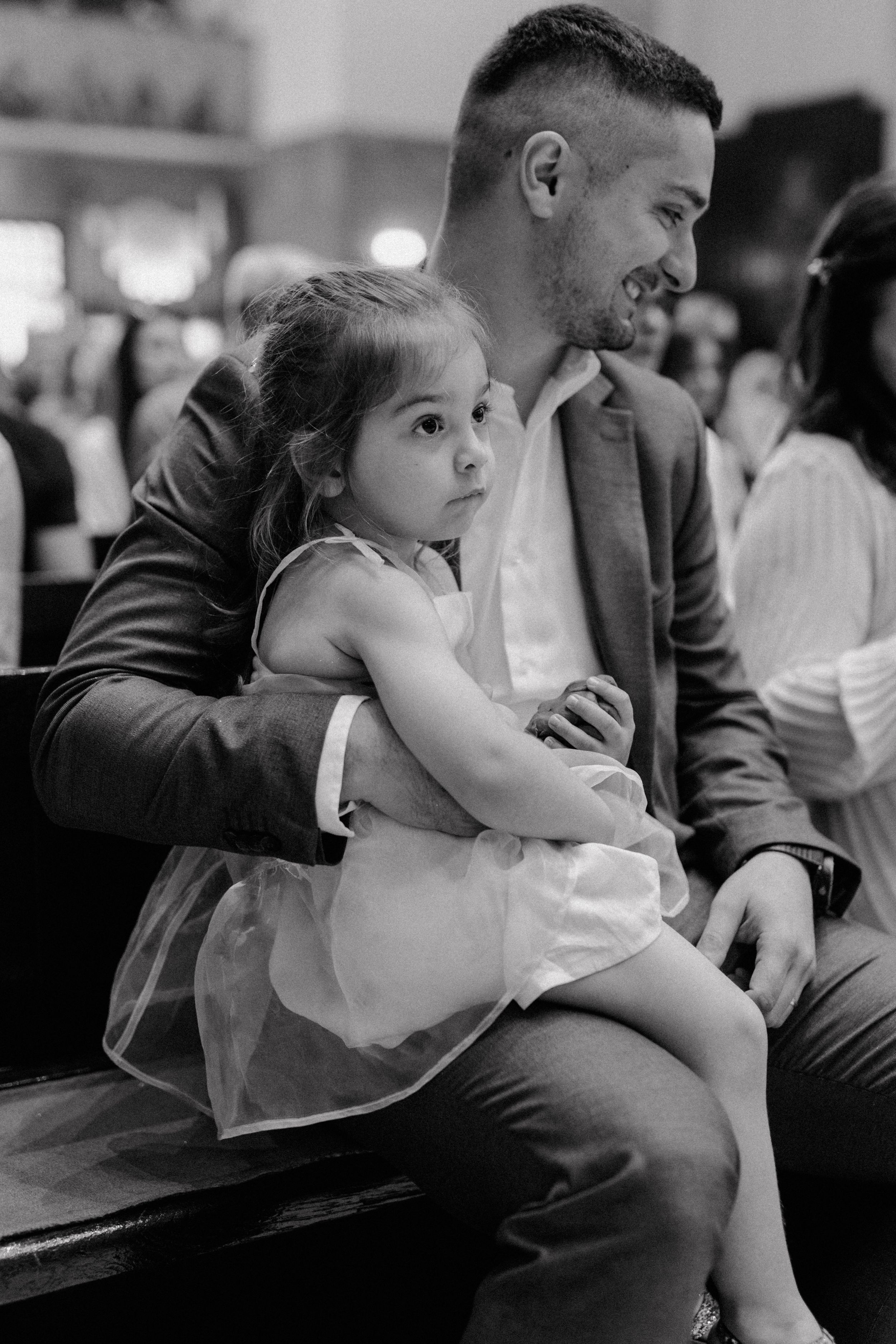 Black and white photo of an older daughter sitting on her dad's lap during the christening ceremony.