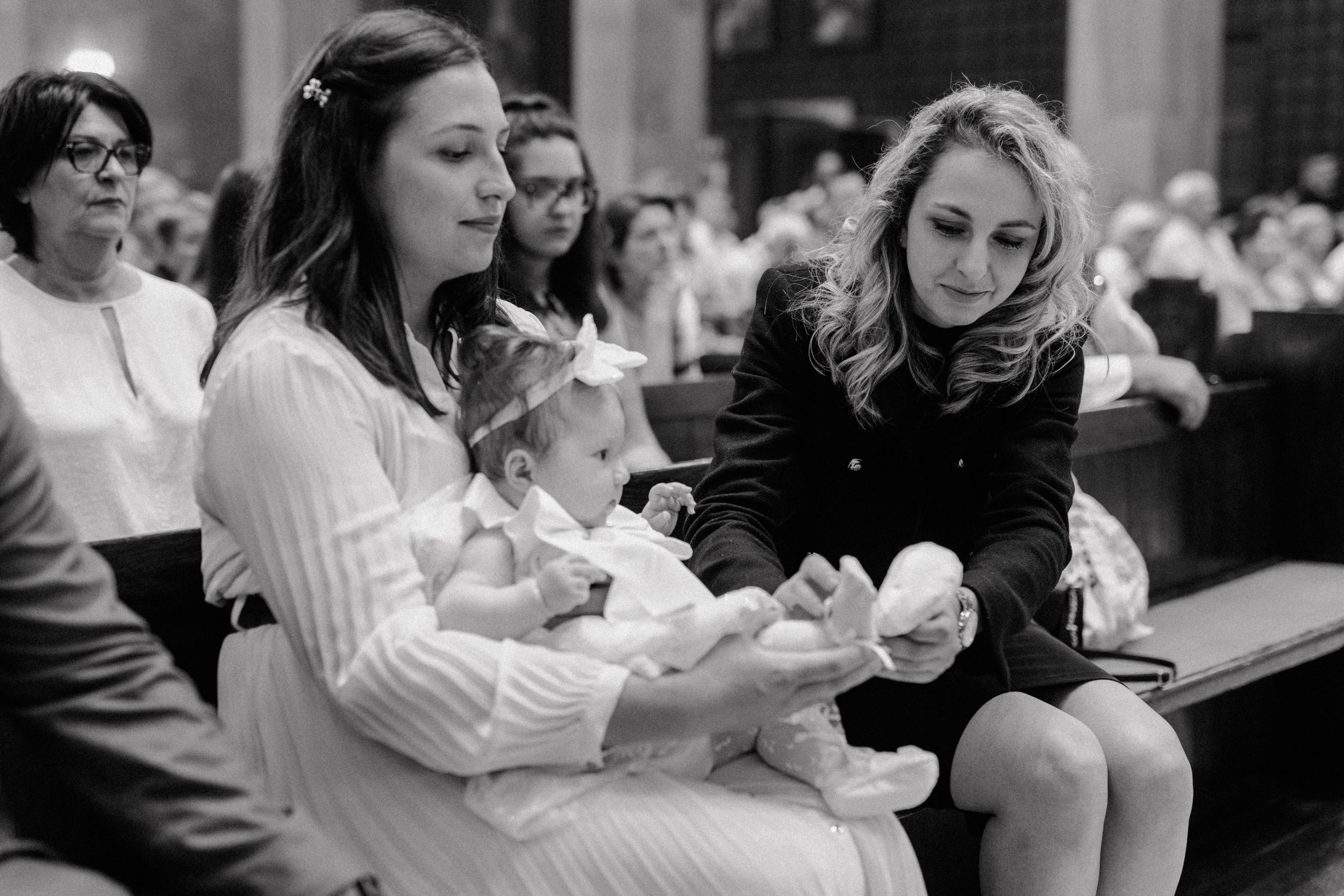 Godmother gently placing a tiny shoe on the baby, who is sitting on mom's lap during the christening ceremony.