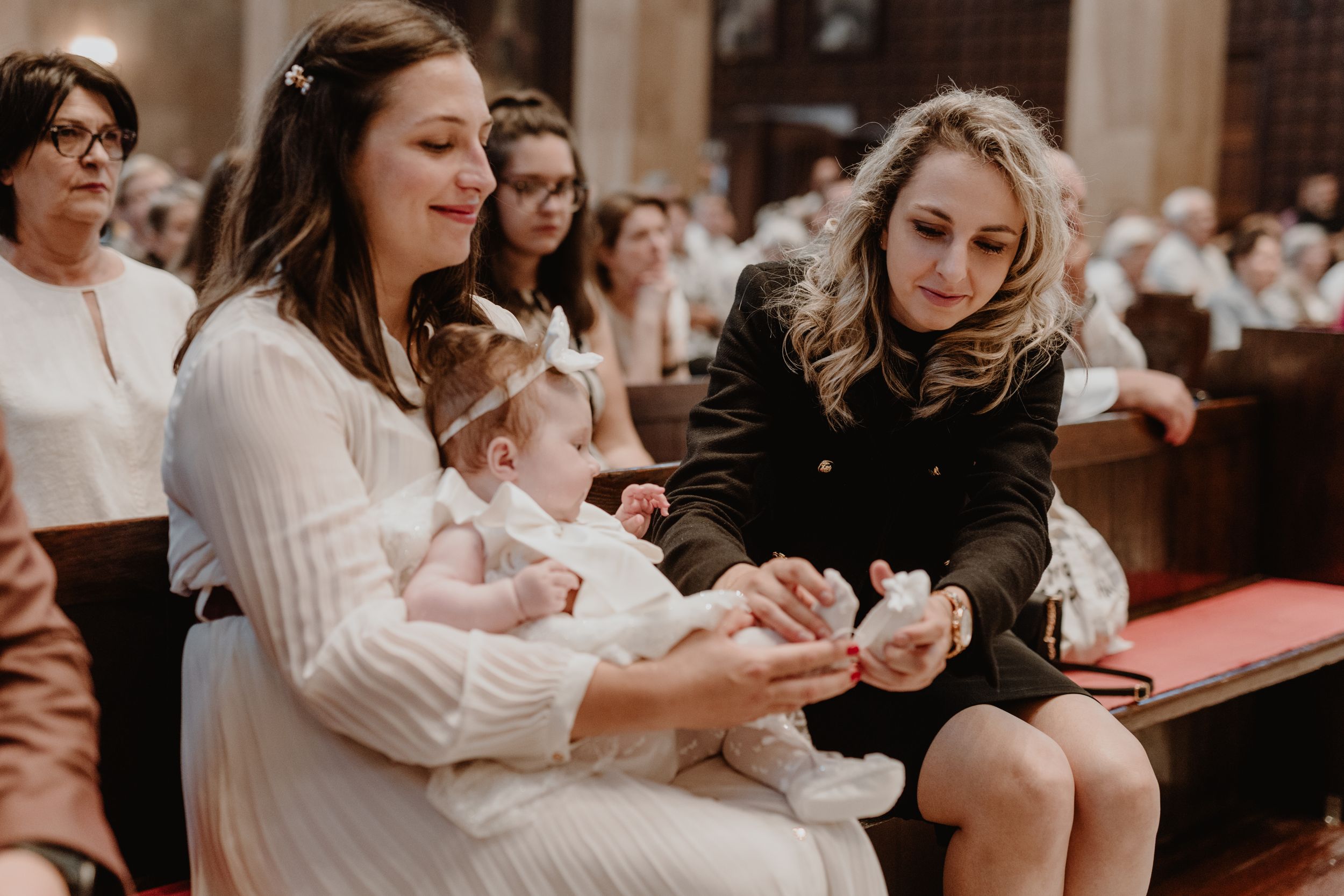 odmother carefully adjusting a tiny shoe on the baby’s foot while the baby sits on mom’s lap,during the christening.