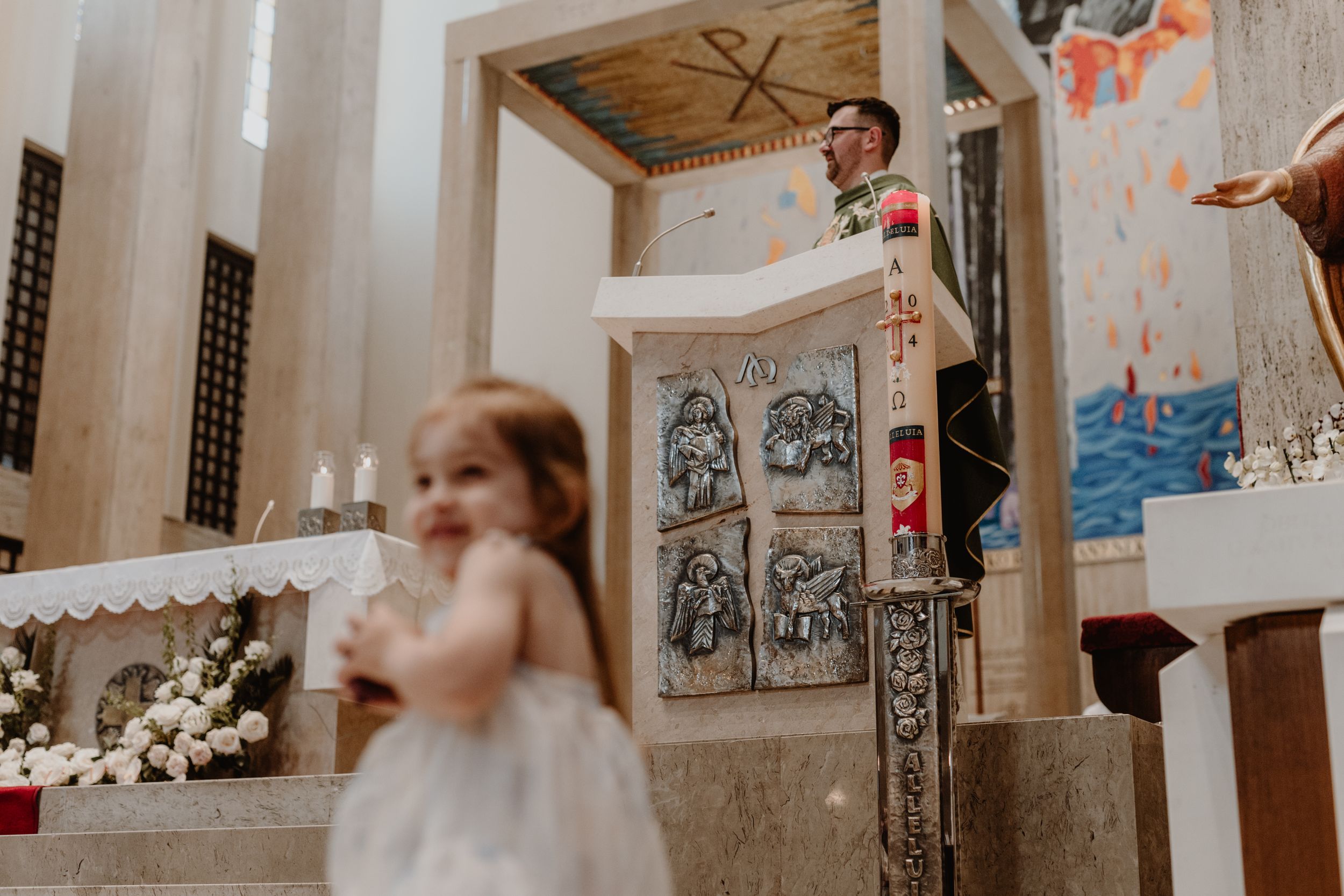 Older daughter in the foreground, slightly out of focus, while the priest visible in the background during christening.