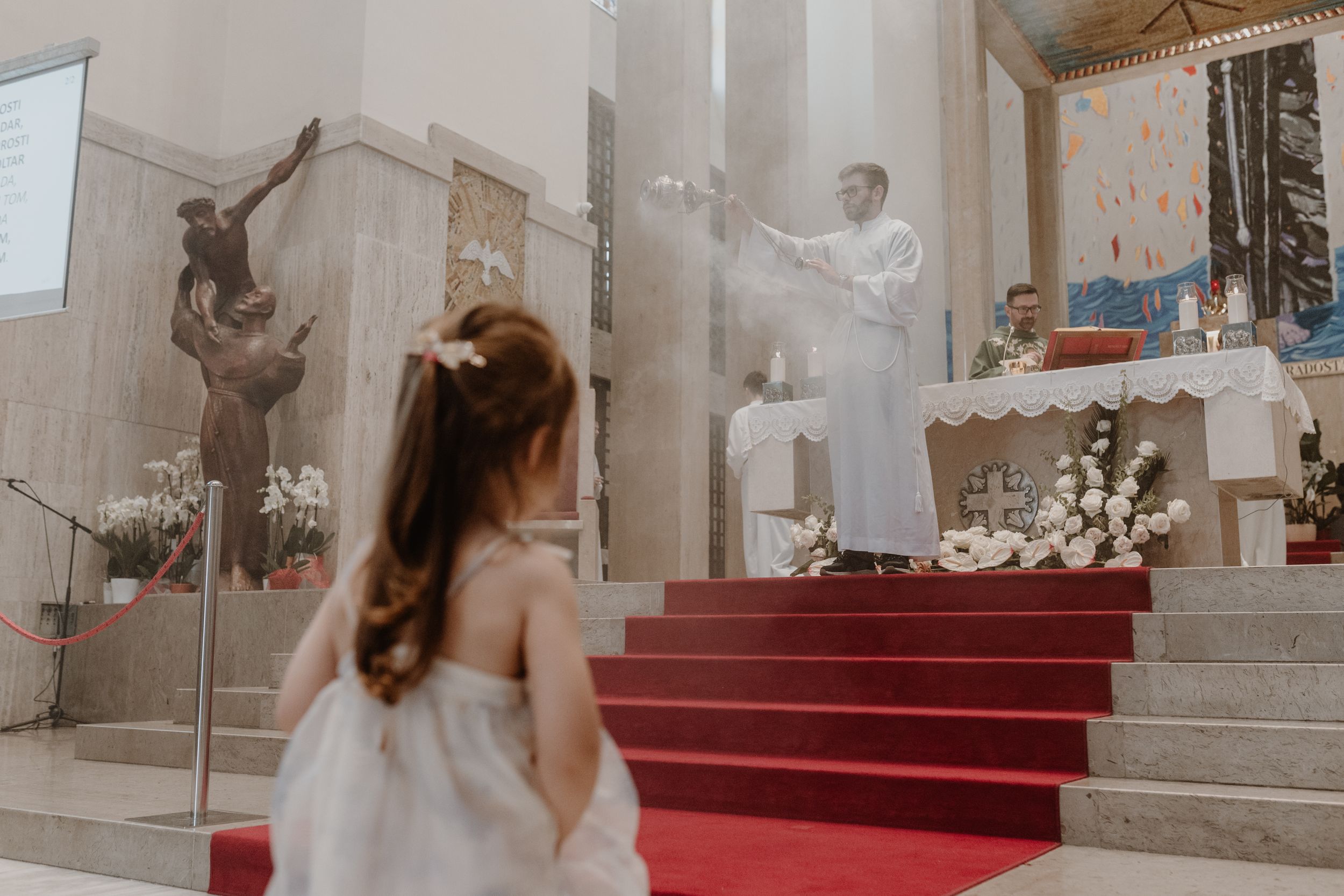 Daughter out of focus in the foreground, while an altar boy spreads incense at the altar in a wide shot.
