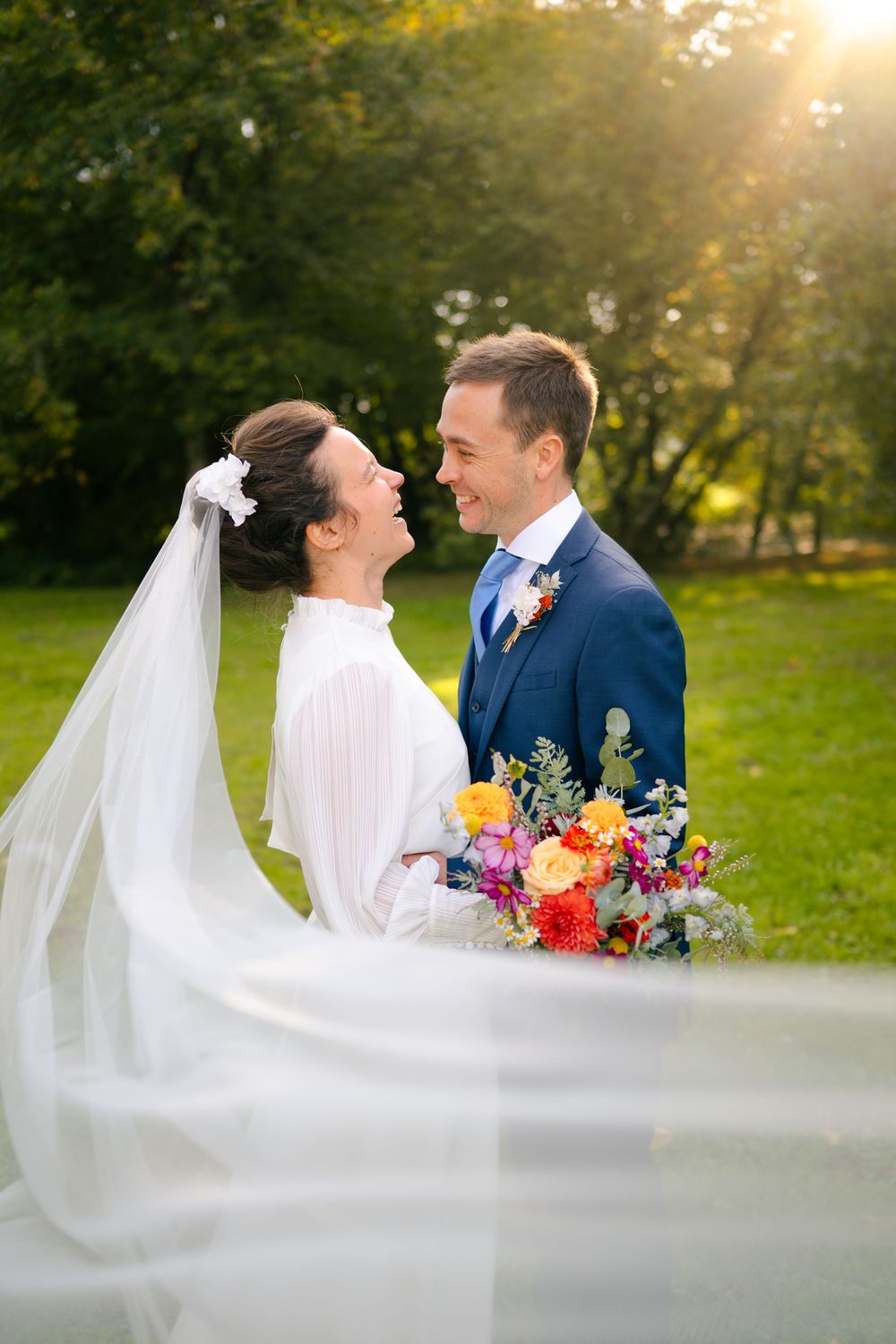 Newlywed couple shares romantic moments in a garden setting at sunset with bride's flowing veil and colorful bouquet.