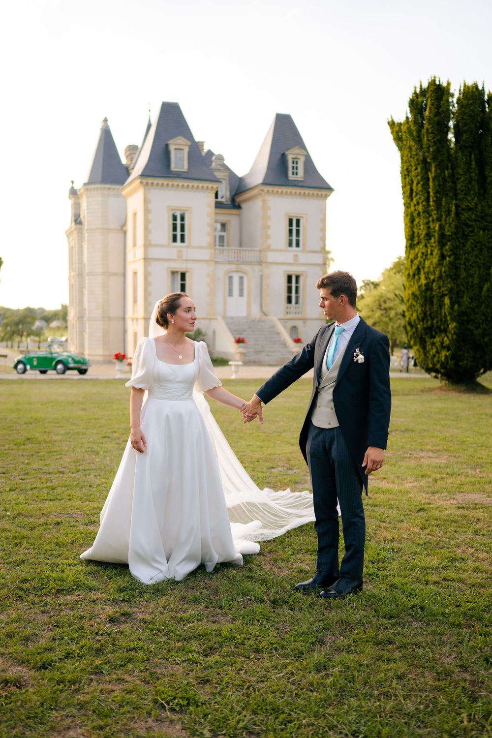 A wedding couple shares a joyful moment on the lawn of a majestic French chateau with elegant towers and architecture.