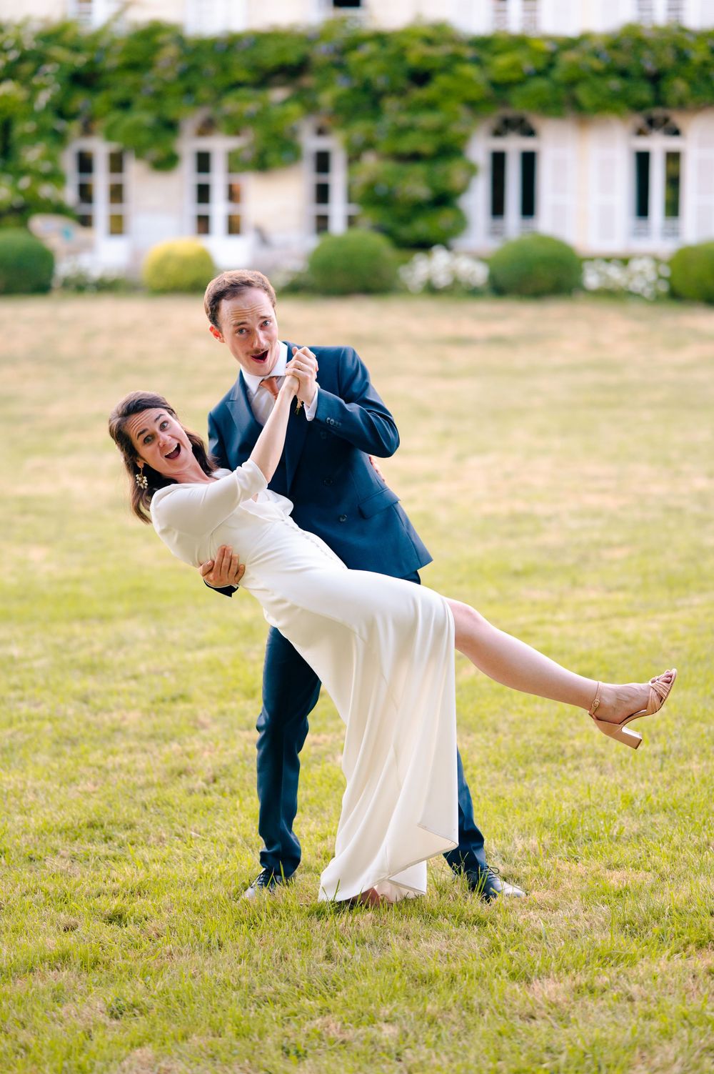 A couple performs a playful dance dip sequence in a grassy field outside a white building on a sunny day.