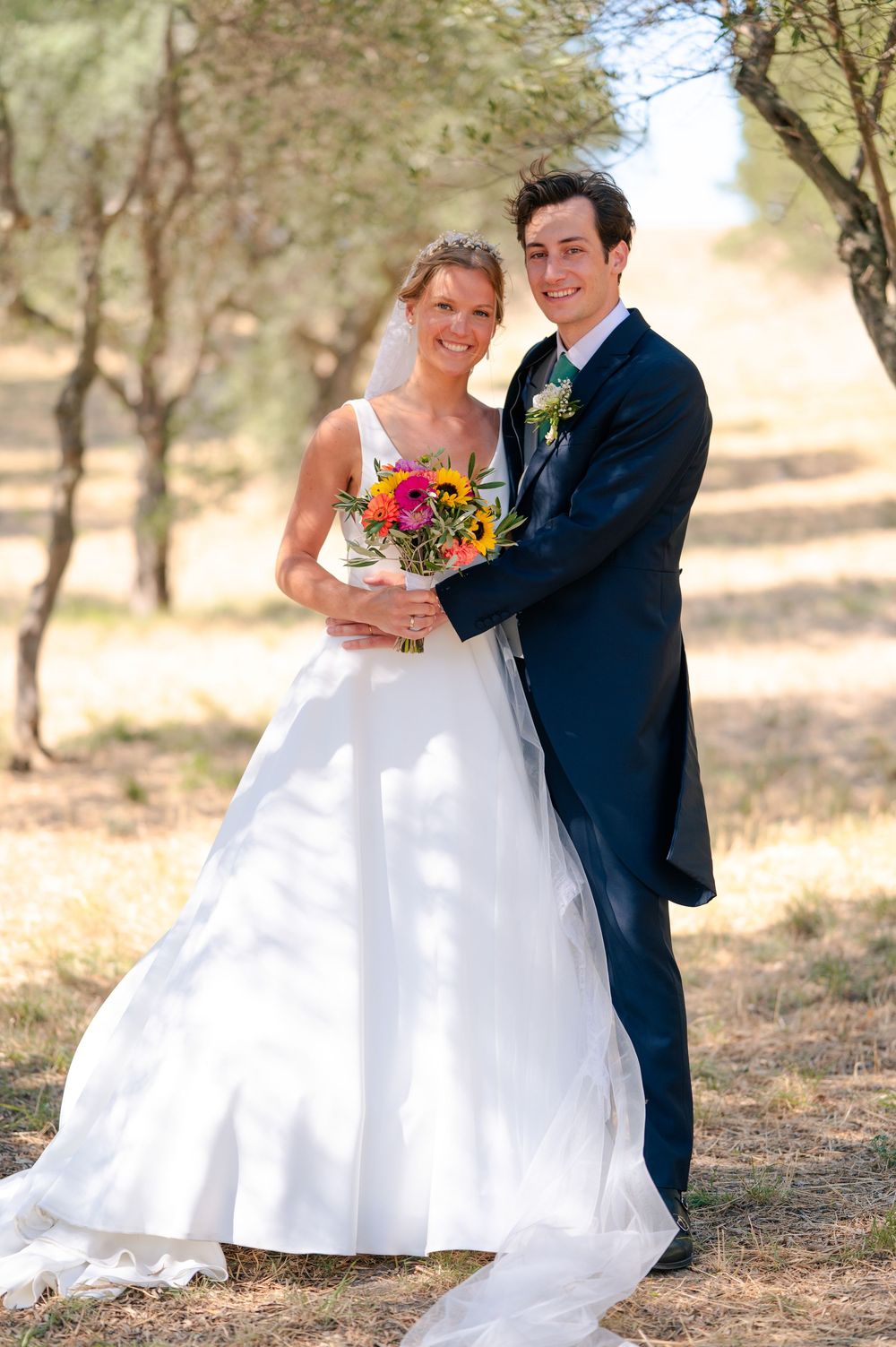 A couple in wedding attire poses together in a rustic outdoor setting with trees and natural lighting.