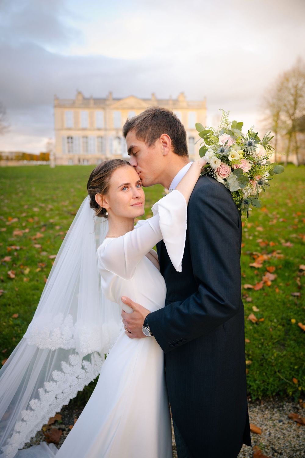 A romantic wedding couple embraces on the lawn of an elegant chateau with autumn leaves scattered on the ground.