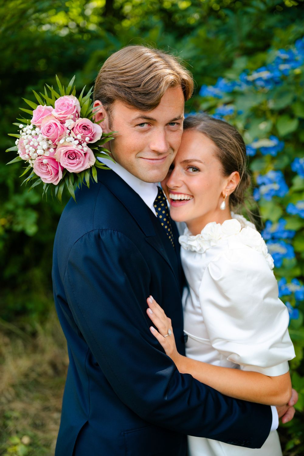 A couple laughs and embraces during their outdoor wedding photoshoot among lush greenery decor and pink floral accents.