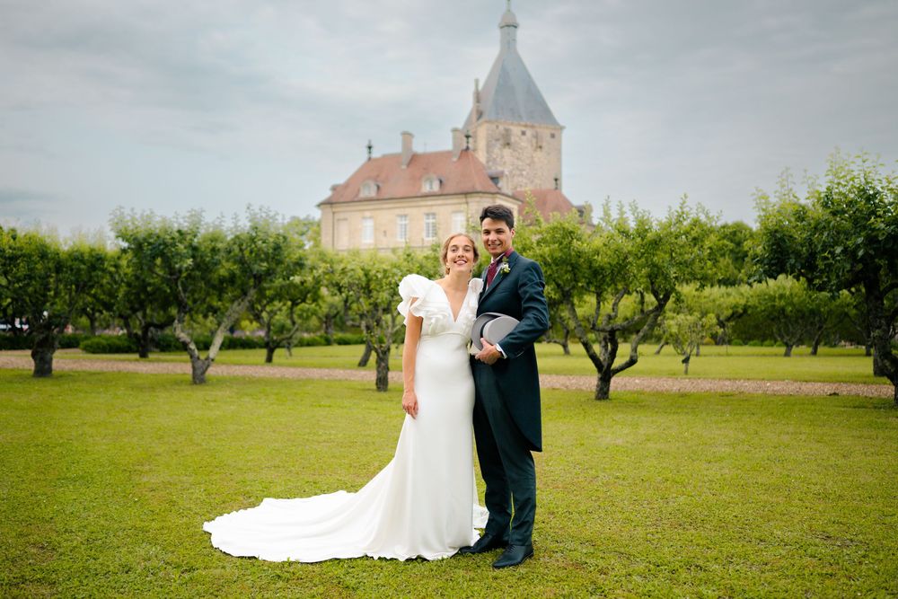 A couple poses in a scenic orchard setting with a historic church steeple visible in the background.