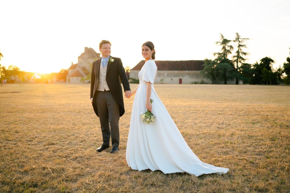 A couple in wedding attire stands in a golden field at sunset, with the bride holding a small bouquet.