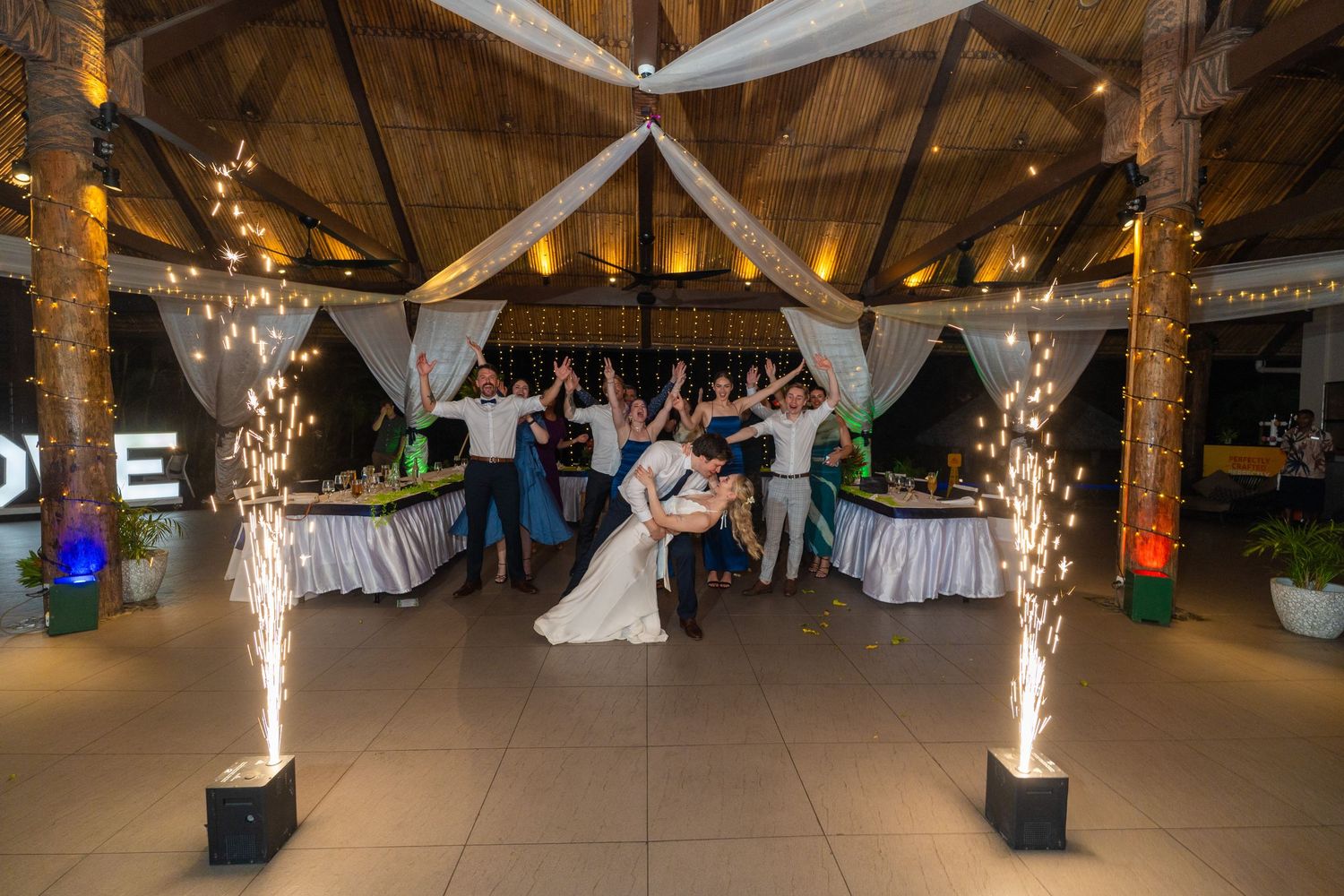 A romantic dip kiss is captured between newlyweds with sparkling firework fountains illuminating their outdoor celebration.