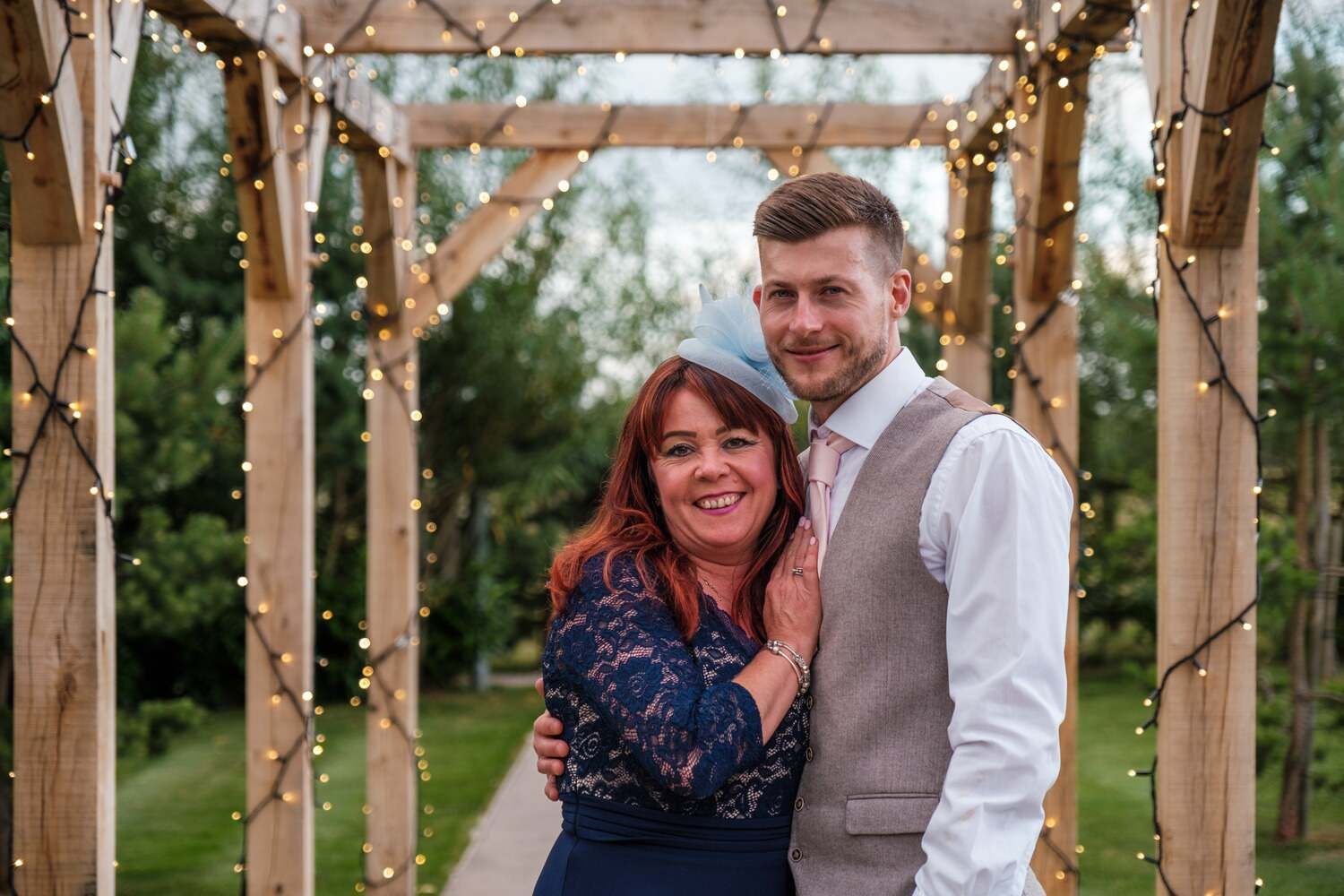 Groom and his mother posing under a wooden arch with string lights.