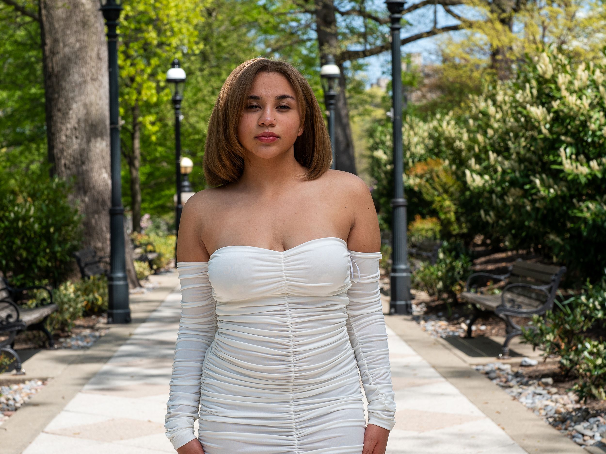 A person in an elegant white ruched dress poses on a park path with lamp posts and greenery.