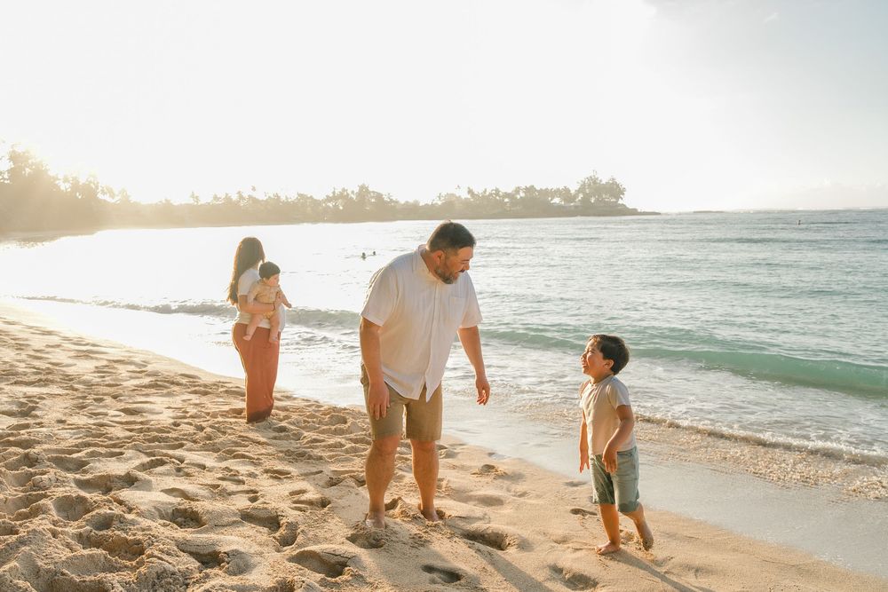 A family walks along a scenic beach at sunset with palm trees and ocean waves in the background.