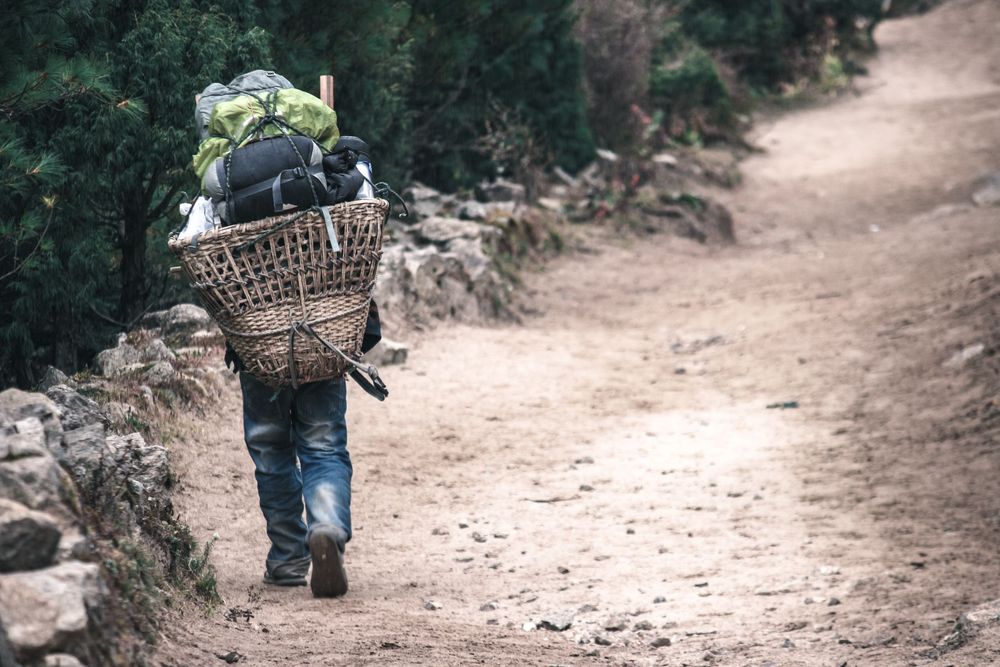 Person carrying a traditional woven basket walks along a dusty mountain trail in a rural setting.