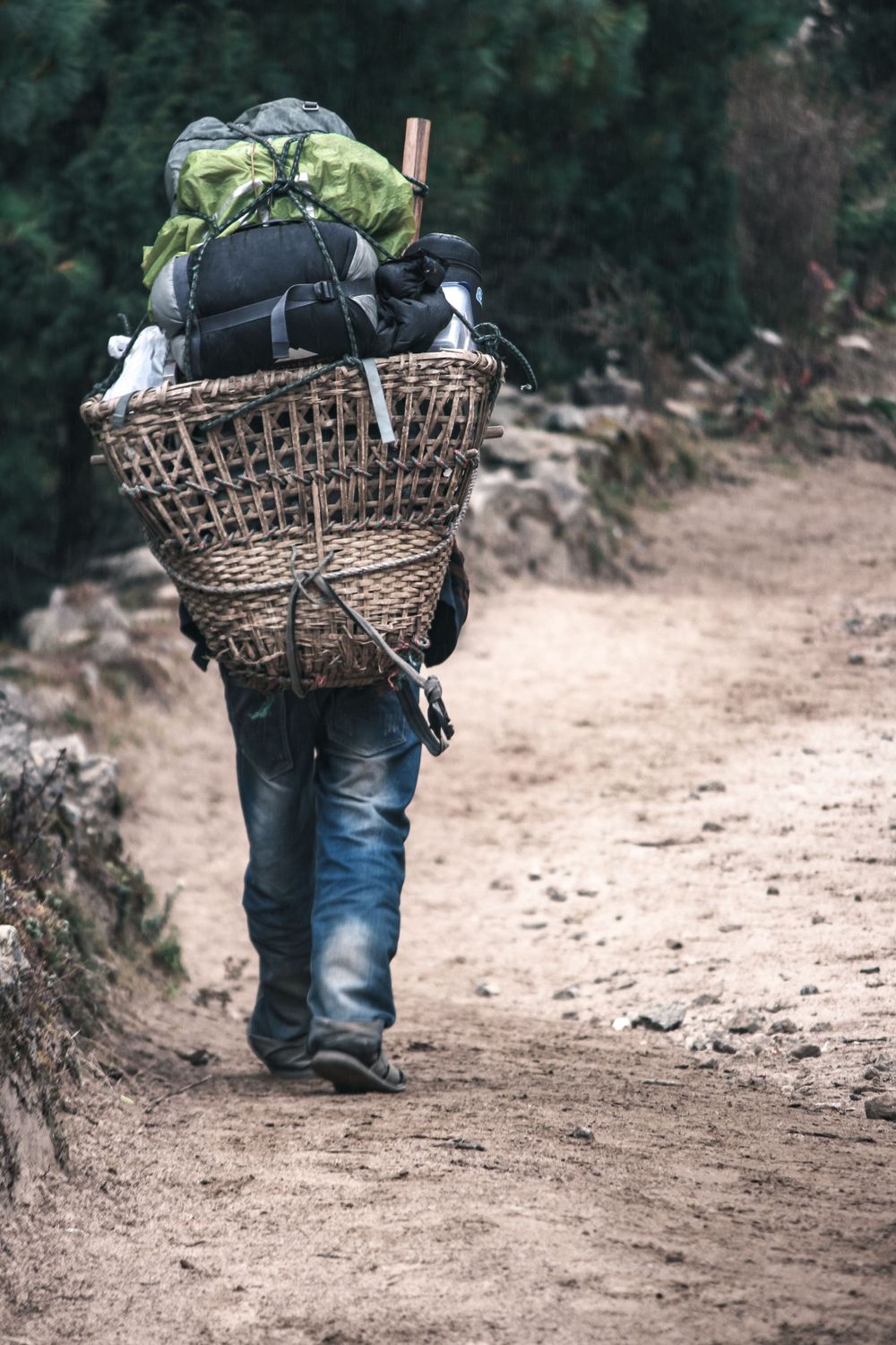Person carrying a woven basket full of supplies walks along a dirt path through wooded terrain.