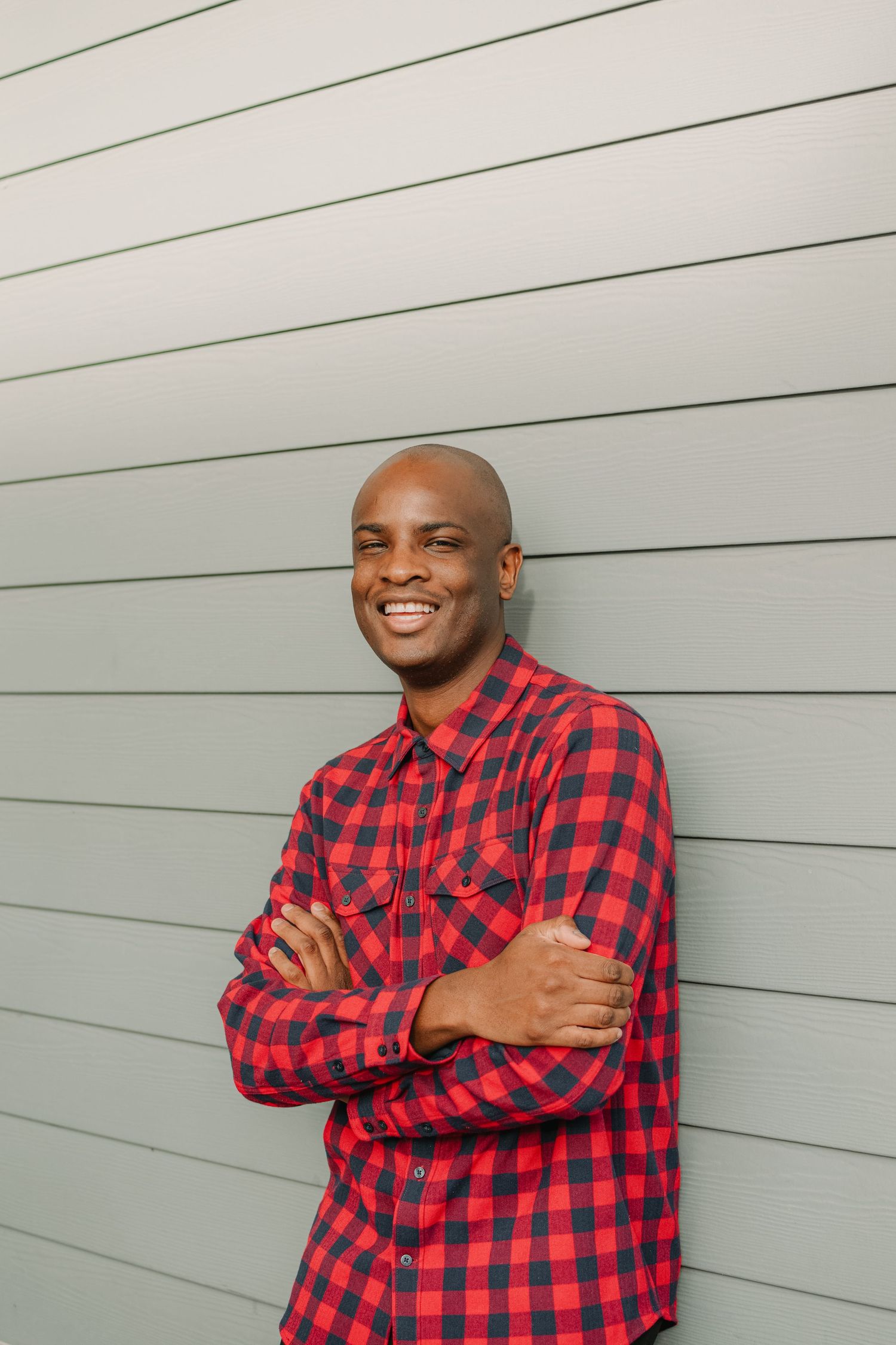 A person in a red plaid shirt stands confidently against a gray striped wall.